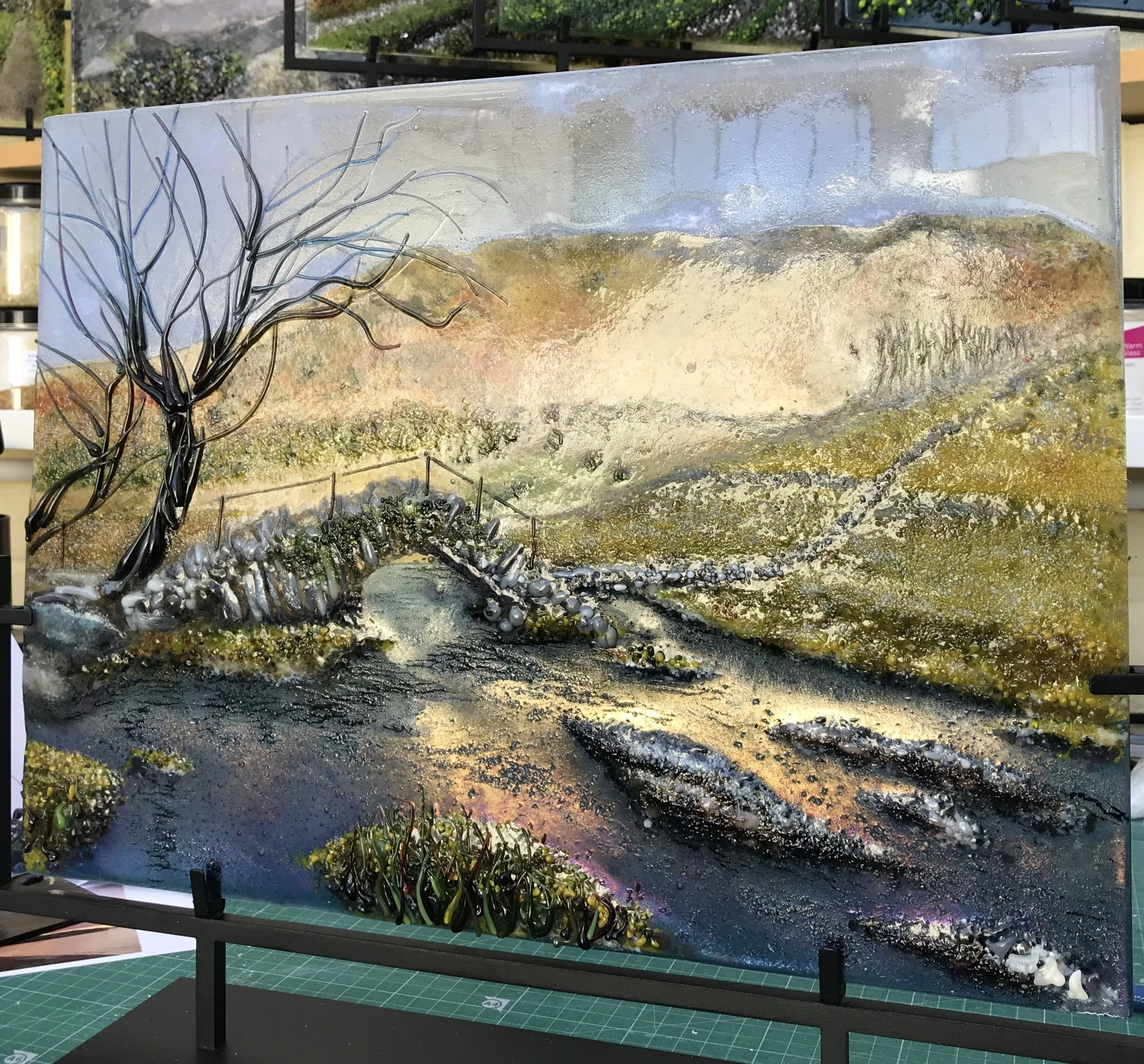 Textured mixed-media landscape painting of a windswept tree, rolling hills, rocky terrain, and a winding stream.