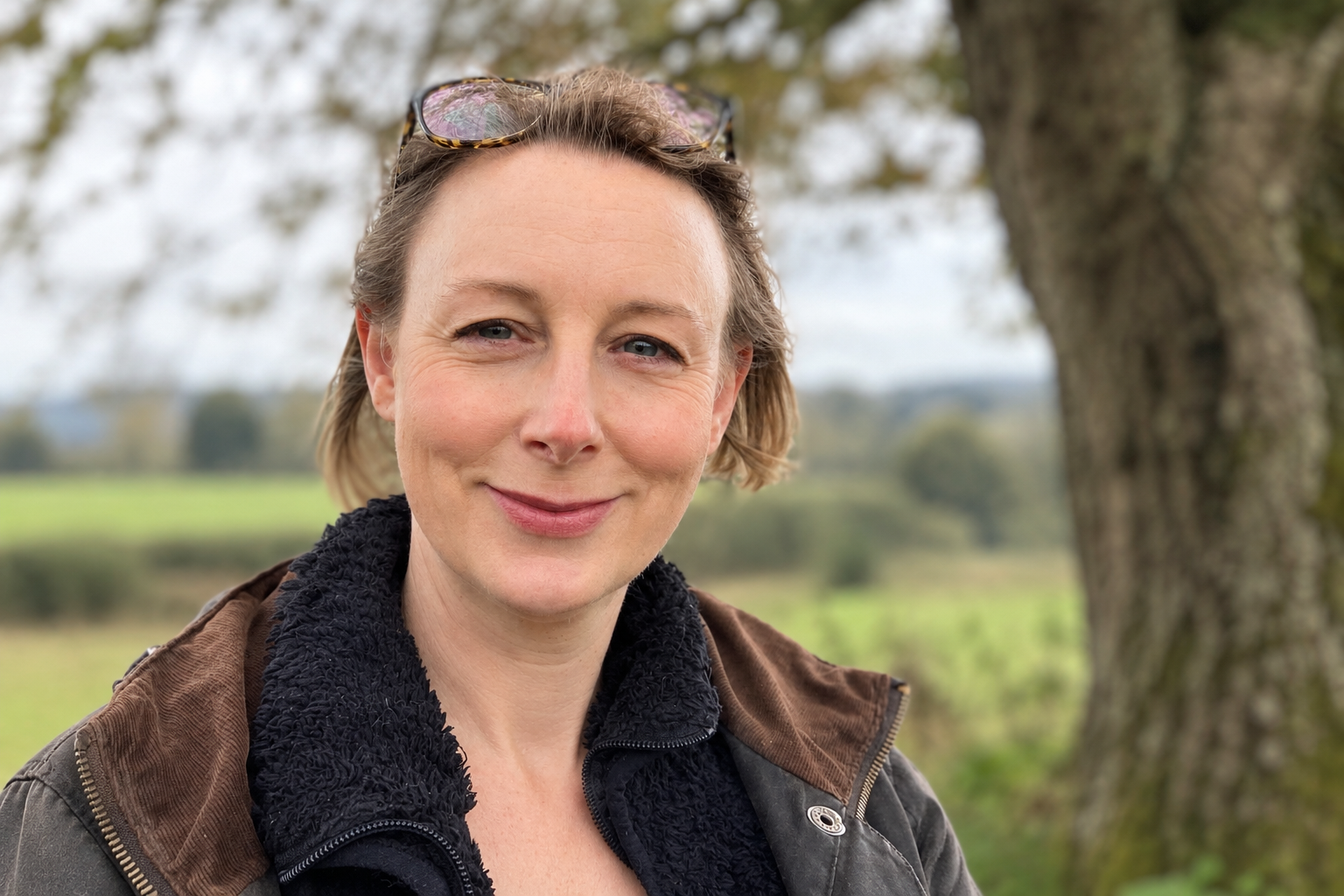 Smiling woman wearing glasses on her head and a fleece-lined jacket outdoors near a tree