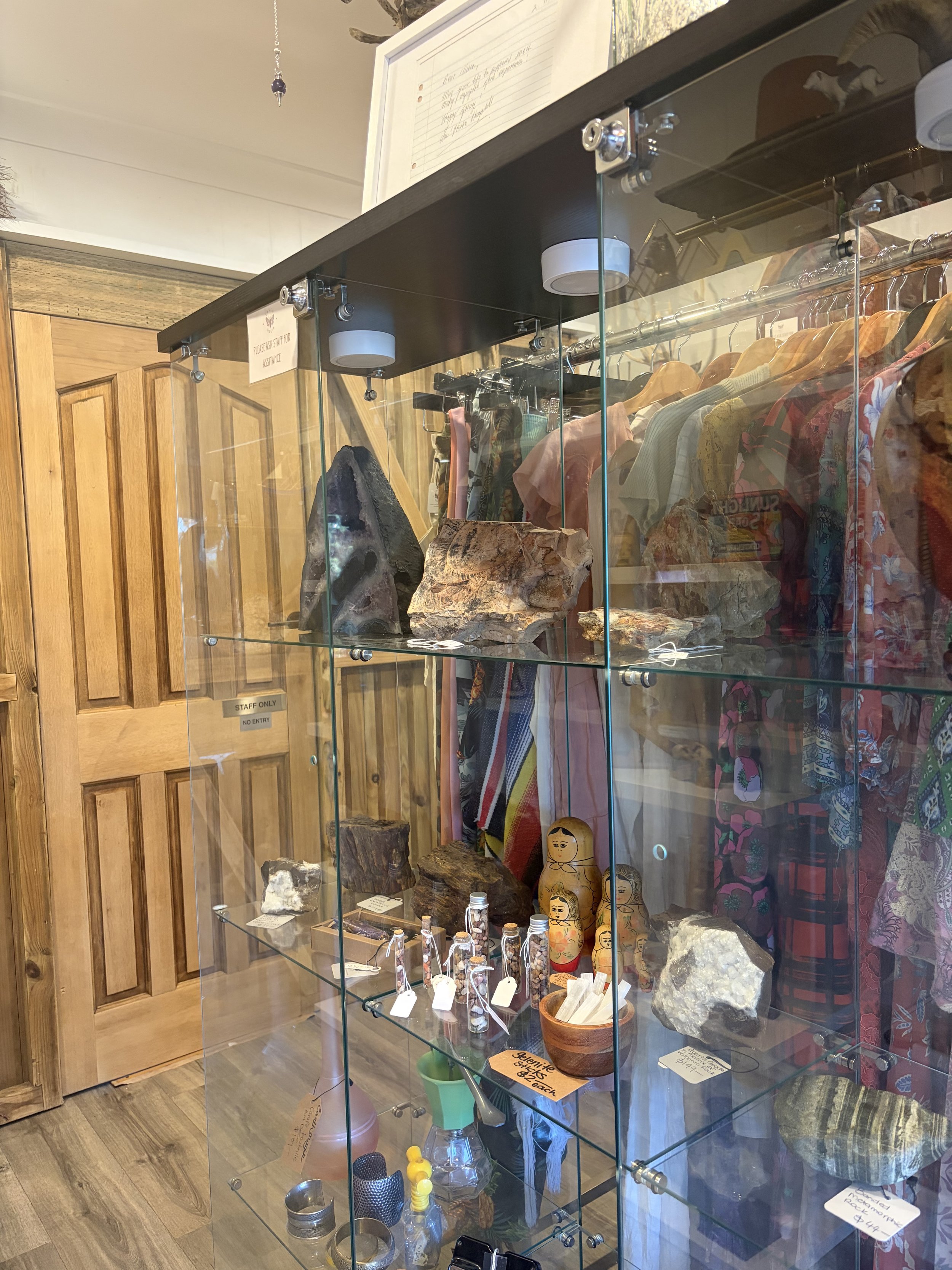 Glass display cabinet filled with rocks, crystals, minerals, handmade dolls, bottles, jewelry, and colorful clothing in a shop.