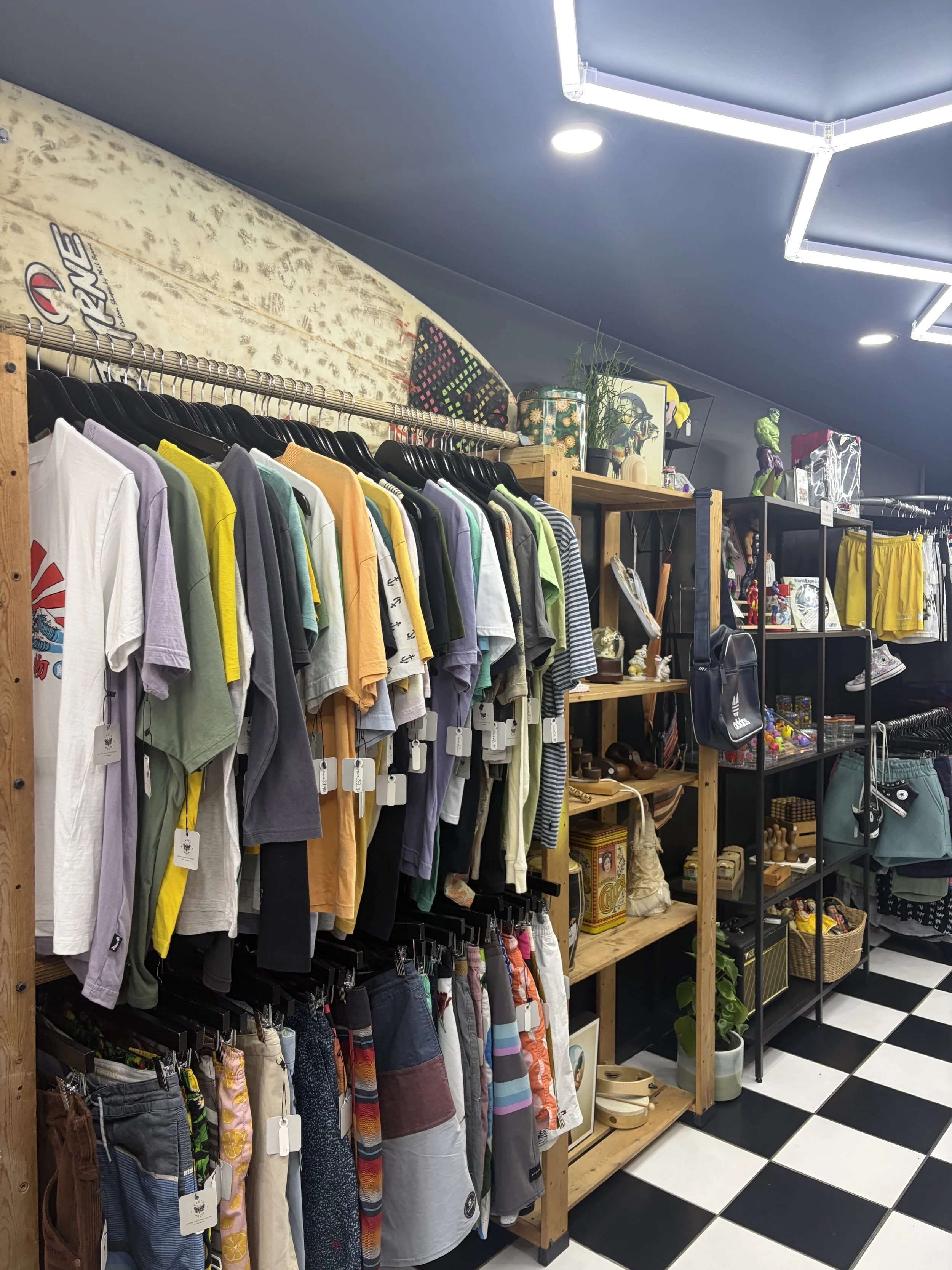 Clothing and gift shop interior with colorful shirts and shorts on racks, shelves of souvenirs and decor, and black-and-white checkered flooring.