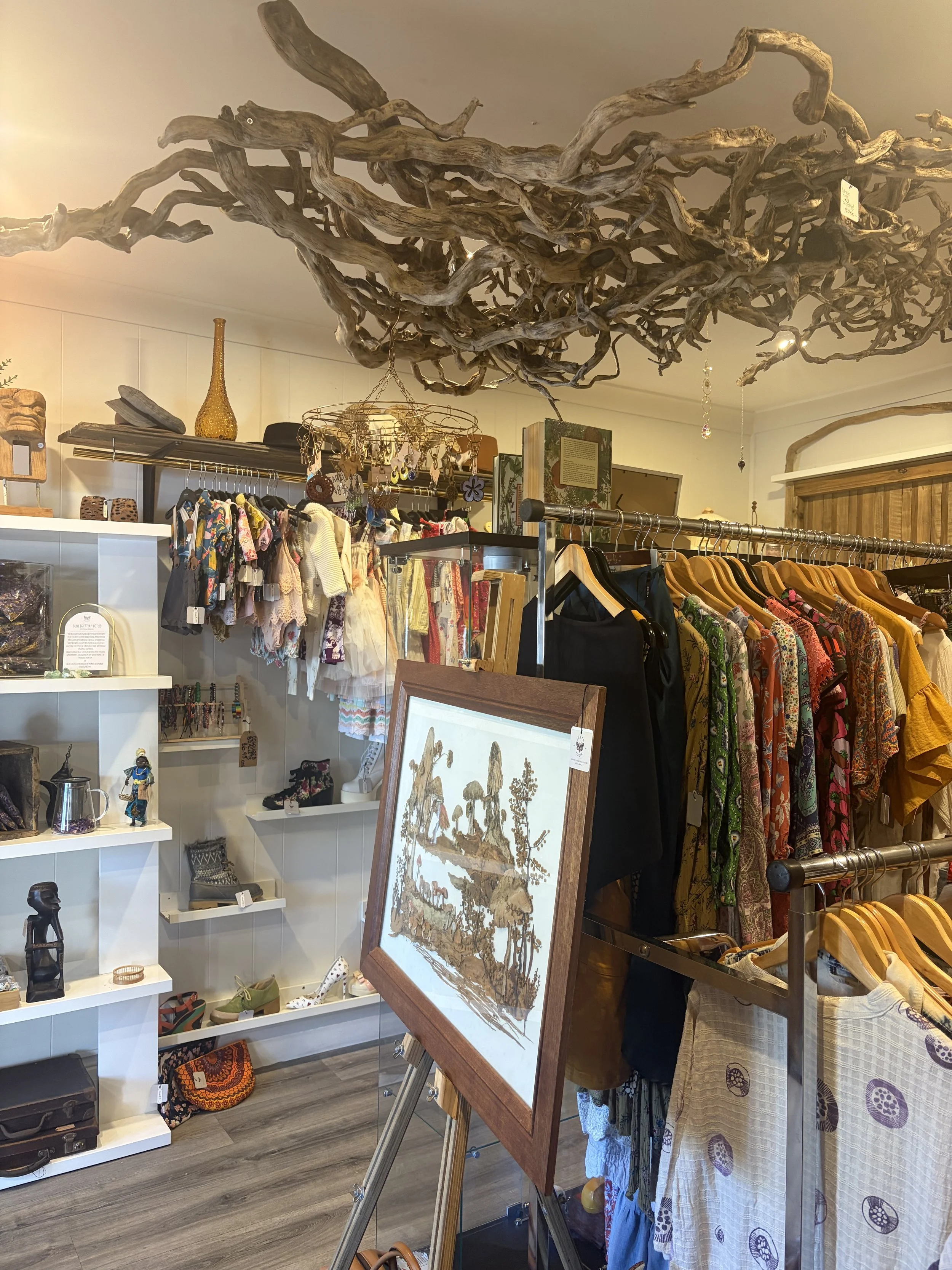 Boutique interior with a large suspended driftwood sculpture, clothing racks, jewelry displays, artwork, shoes, and decorative merchandise.