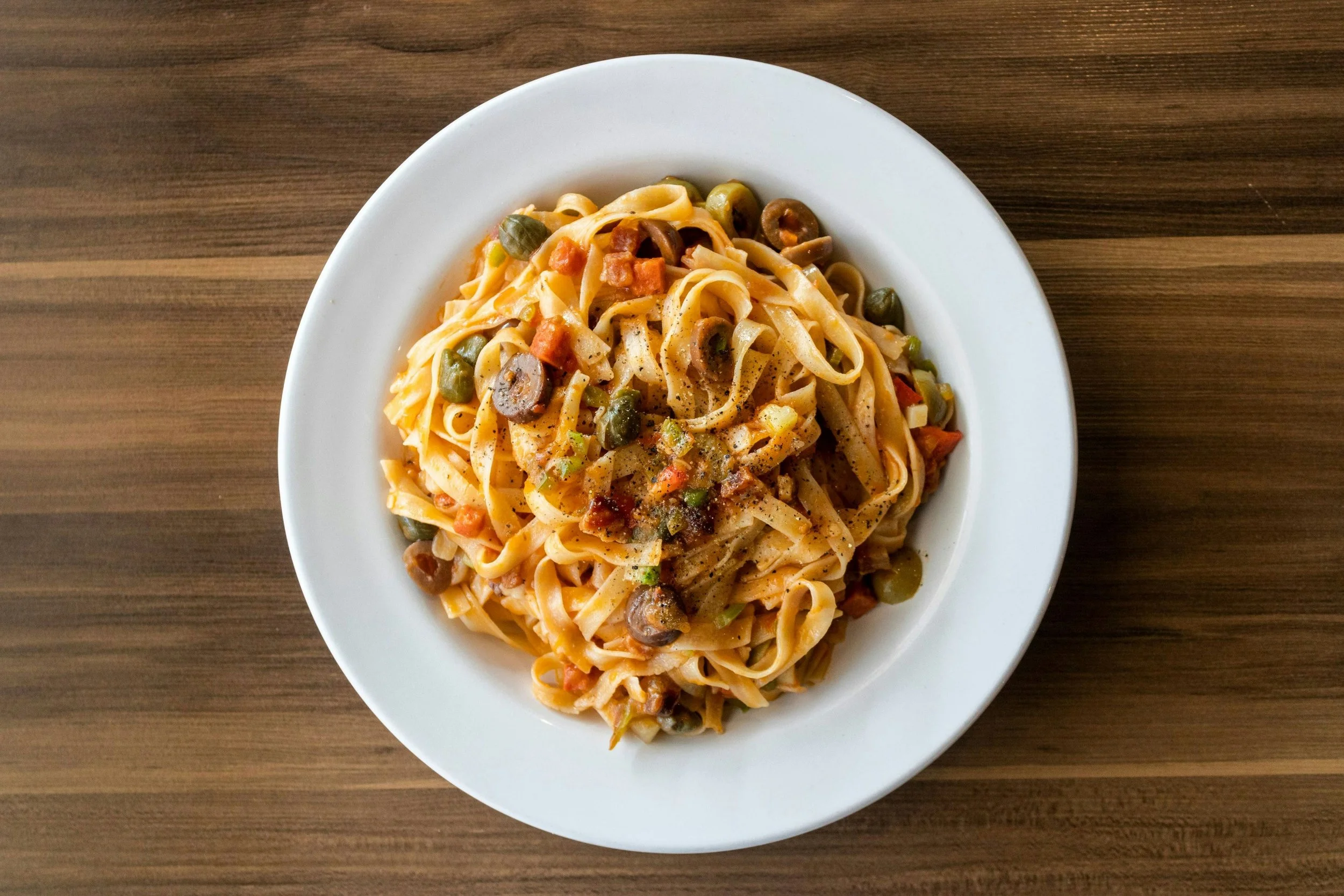 Overhead view of fettuccine pasta with tomato sauce, olives, capers, vegetables, and black pepper on a white plate