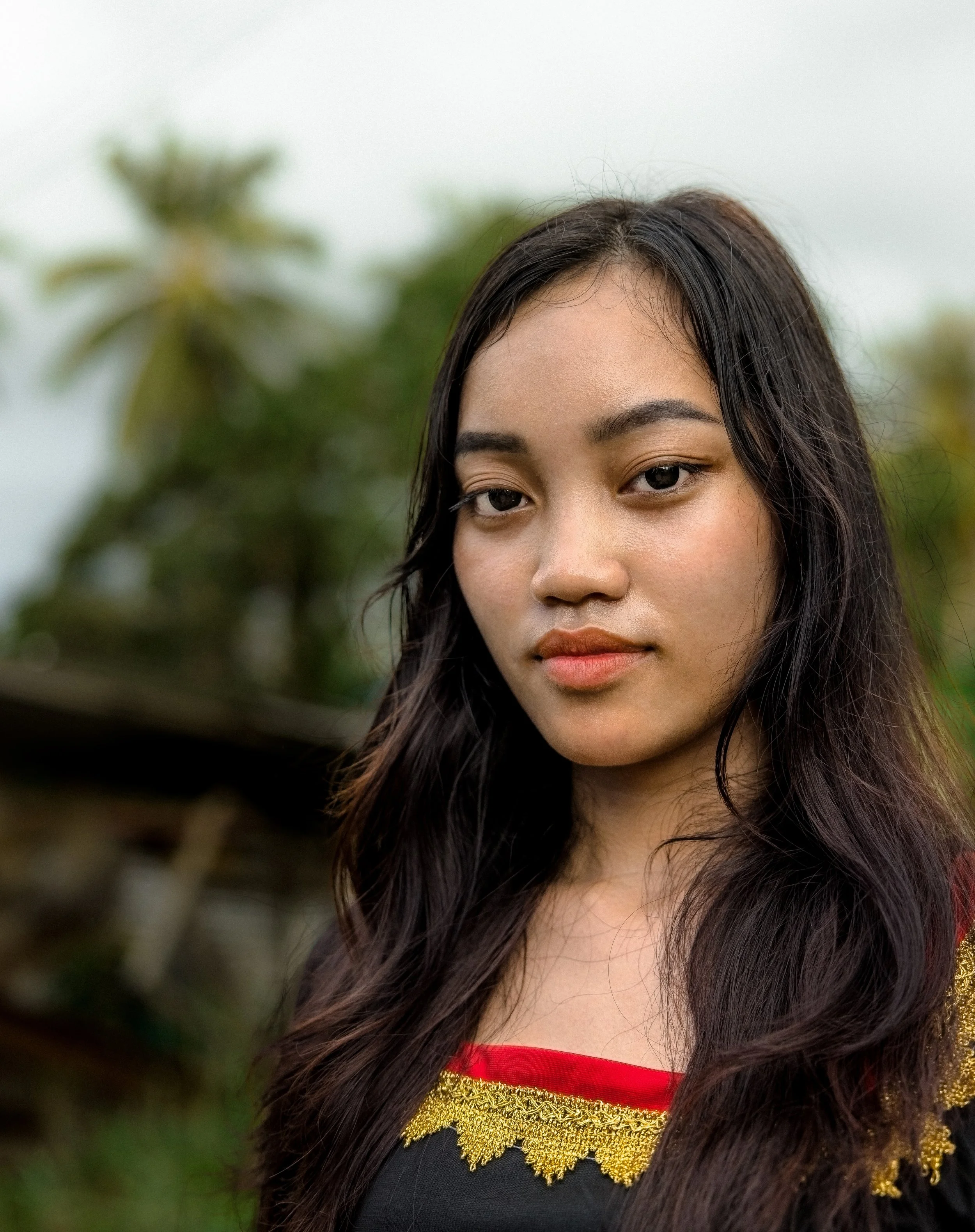 Close-up portrait of a woman in Borneo with long dark hair wearing a black and gold-trimmed outfit outdoors. Photo by David Ferry Jr.