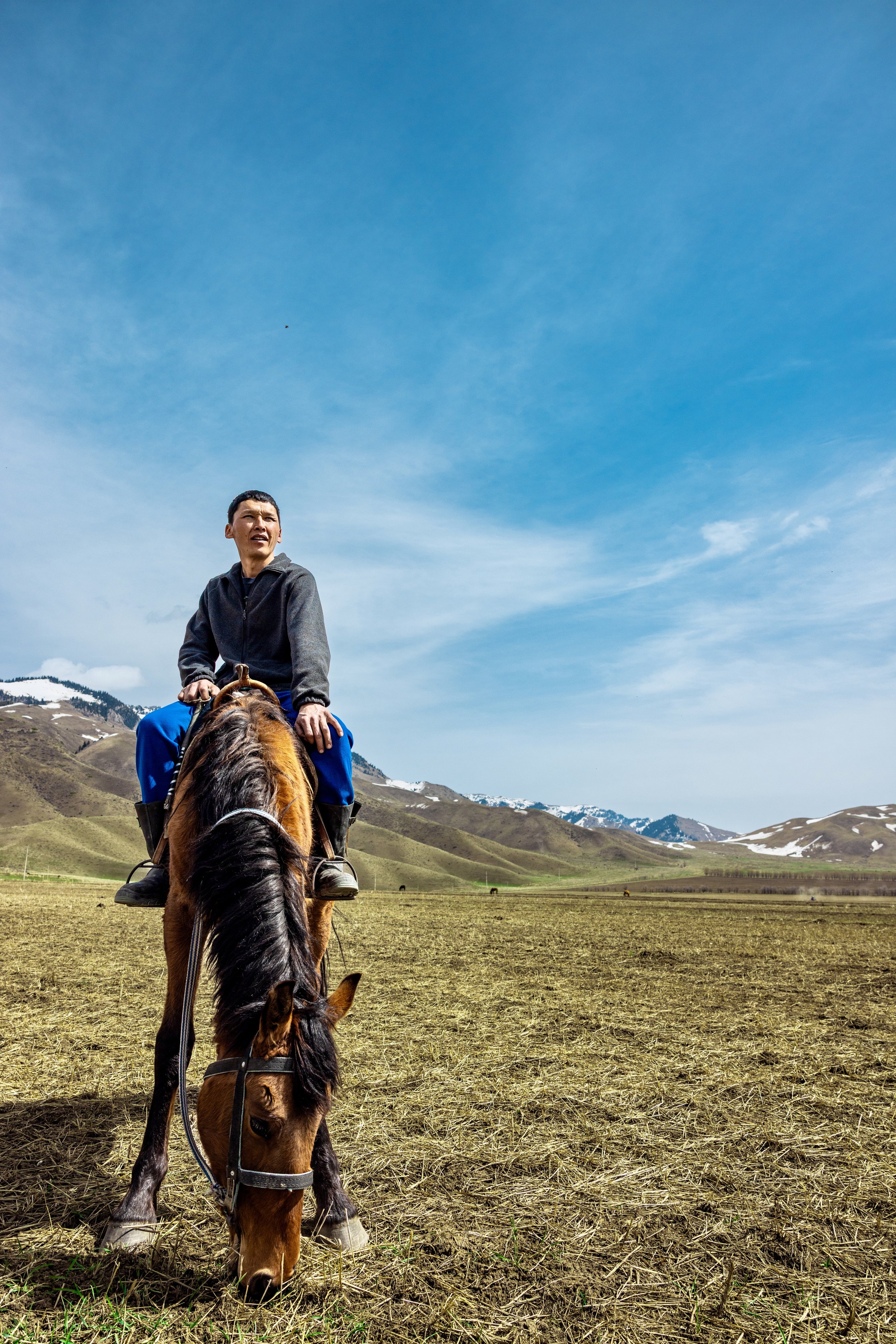 Man riding a horse across a mountain valley with snow-capped peaks under a bright blue sky in Kyrgyzstan. Photo by David Ferry Jr.