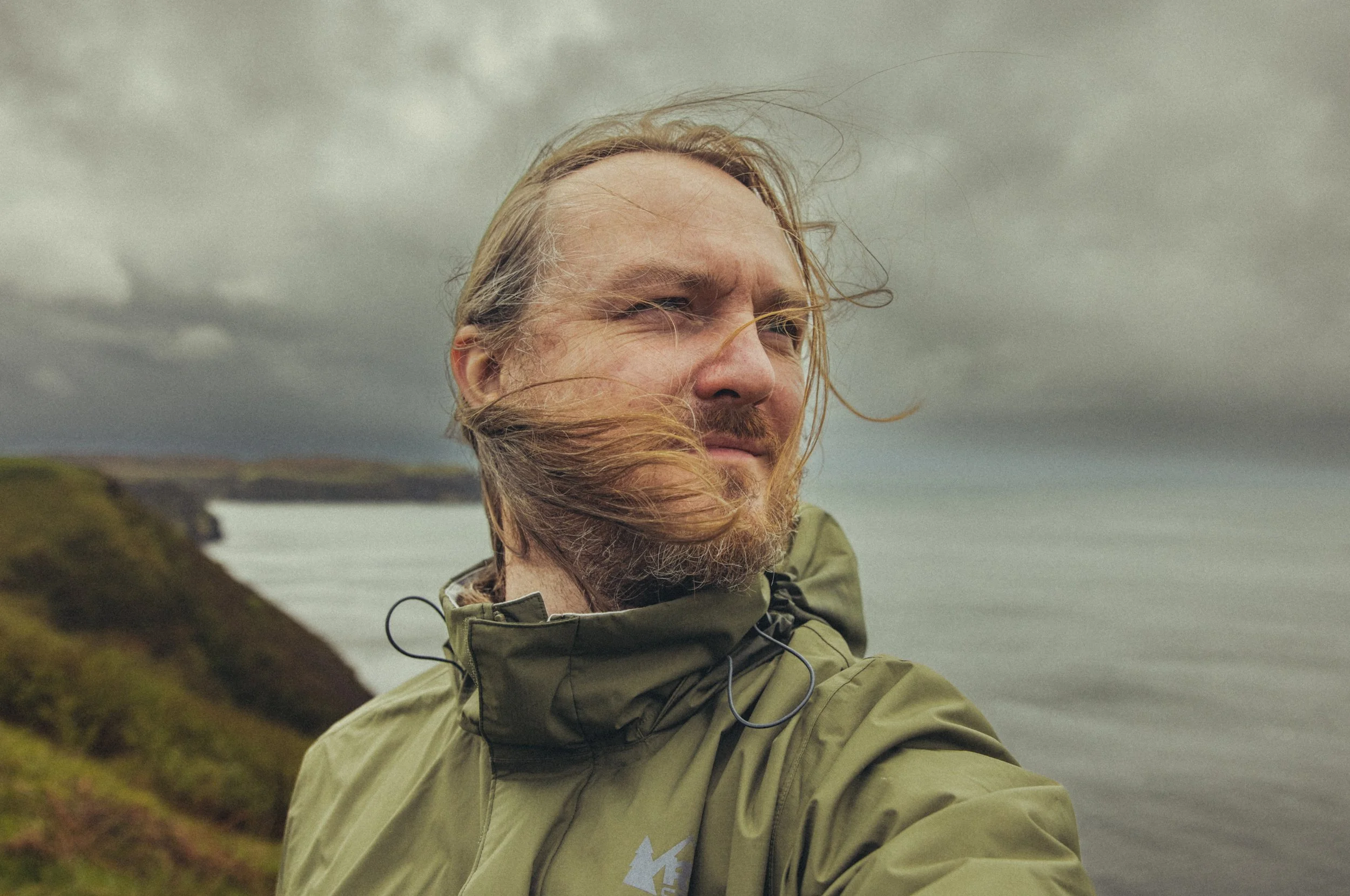 Bearded man in an olive jacket standing outdoors with windblown hair against a misty mountain and lake landscape