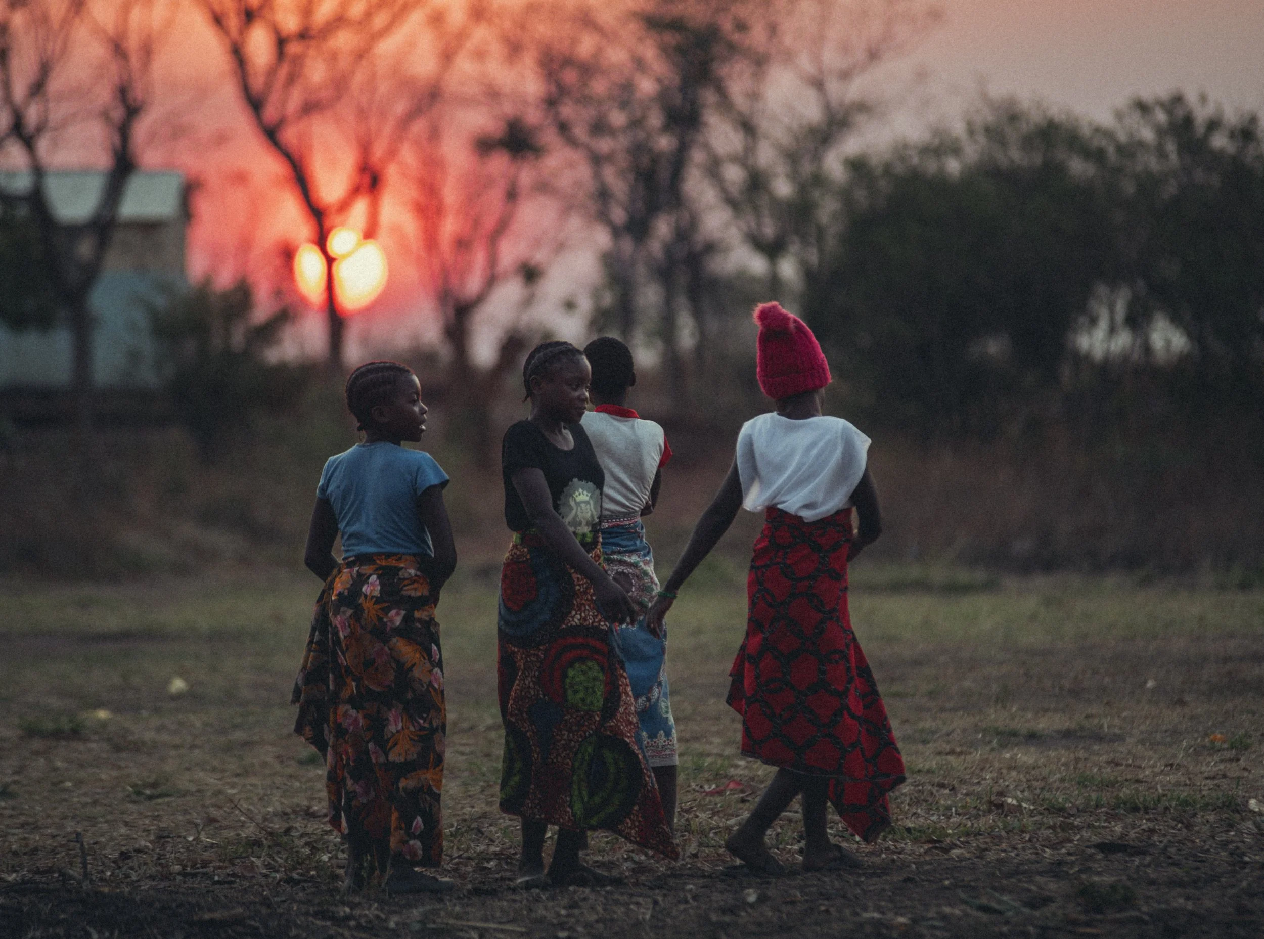 Four Zambian women holding hands and walking outdoors at sunset, wearing colorful patterned skirts and clothing. Photo by David Ferry Jr.