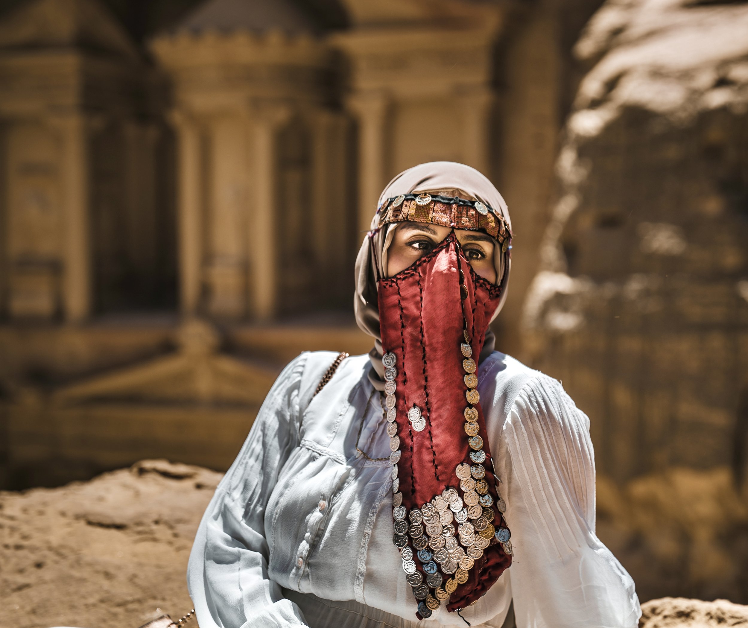 Woman wearing traditional Middle Eastern clothing, a red face veil, headscarf, and coin jewelry in front of sandstone ruins of Petra. Photo by David Ferry Jr.