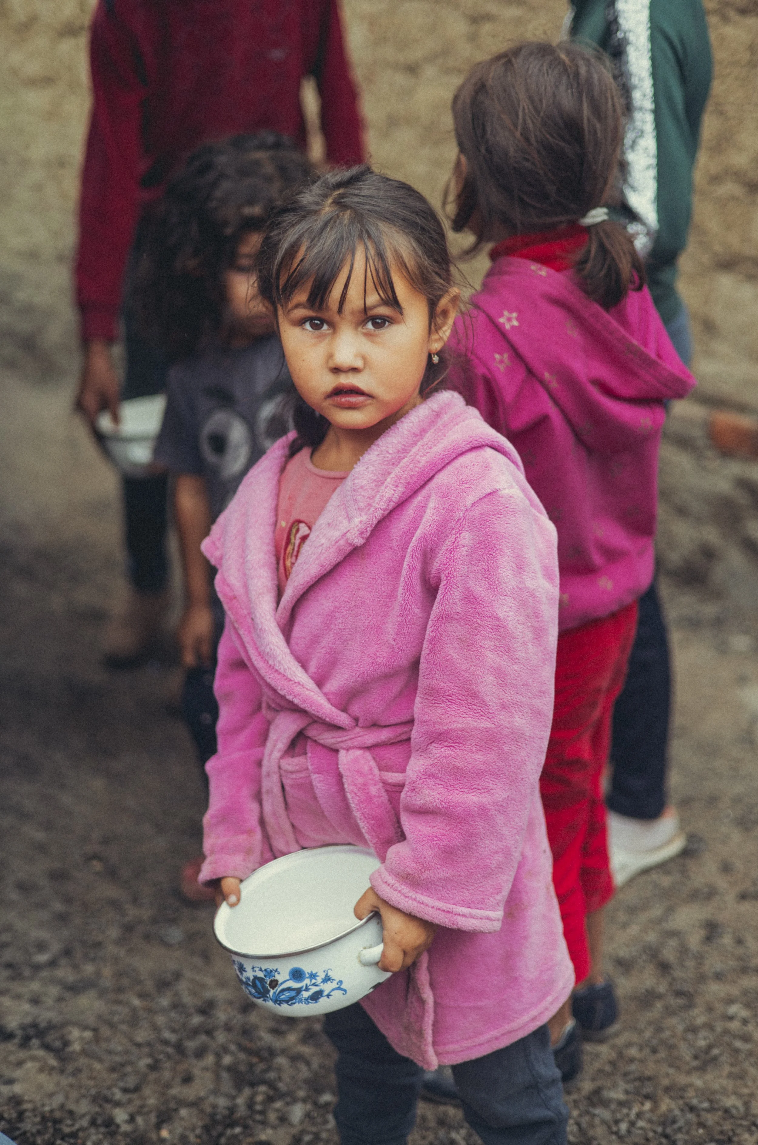 Young Ukrainian girl in a pink fleece jacket holding a floral enamel bowl outdoors, with children in colorful clothing behind her. Photo by David Ferry Jr.
