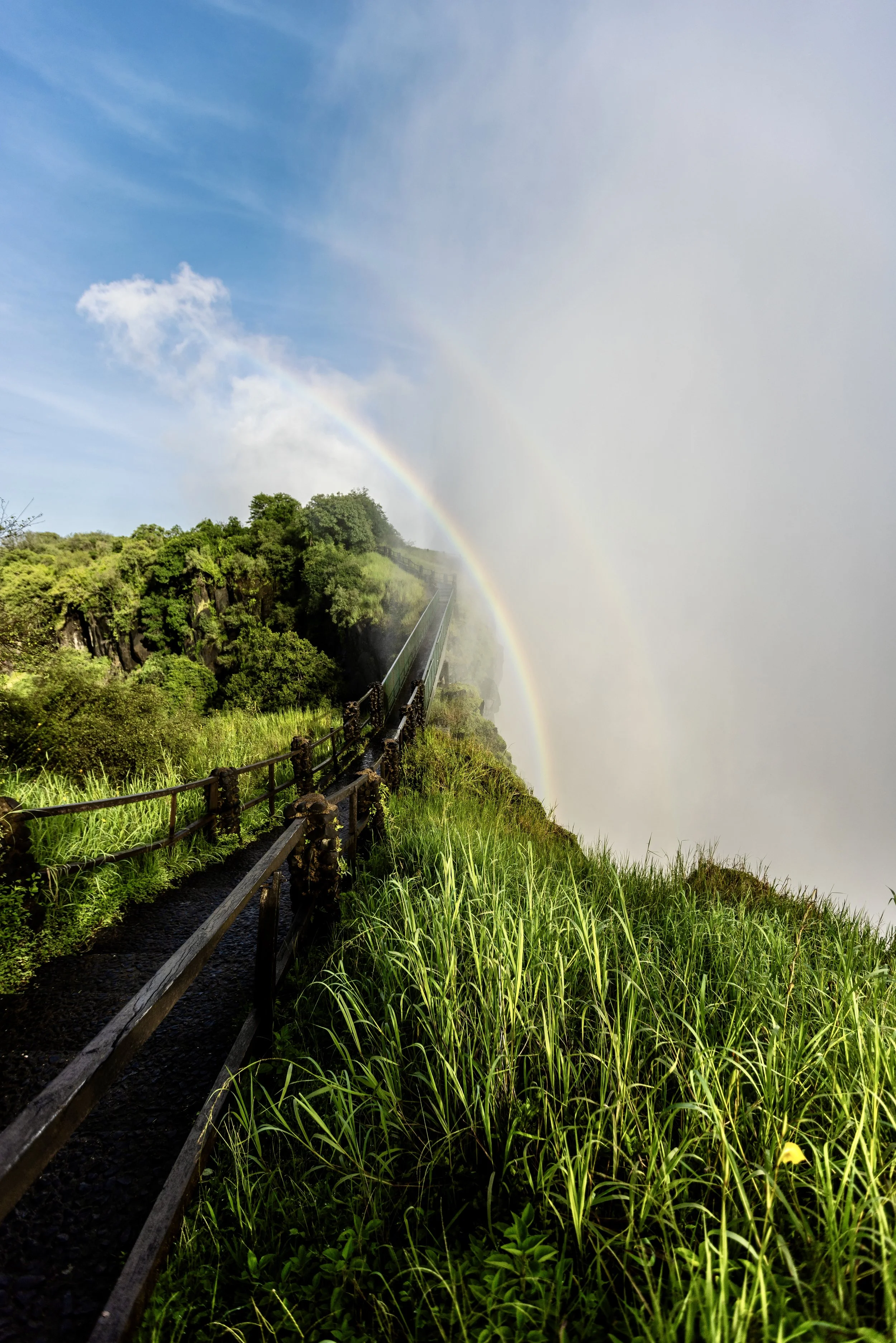 Misty mountain walkway surrounded by lush green vegetation with a rainbow in the spray at Victoria Falls. Photo by David Ferry Jr.