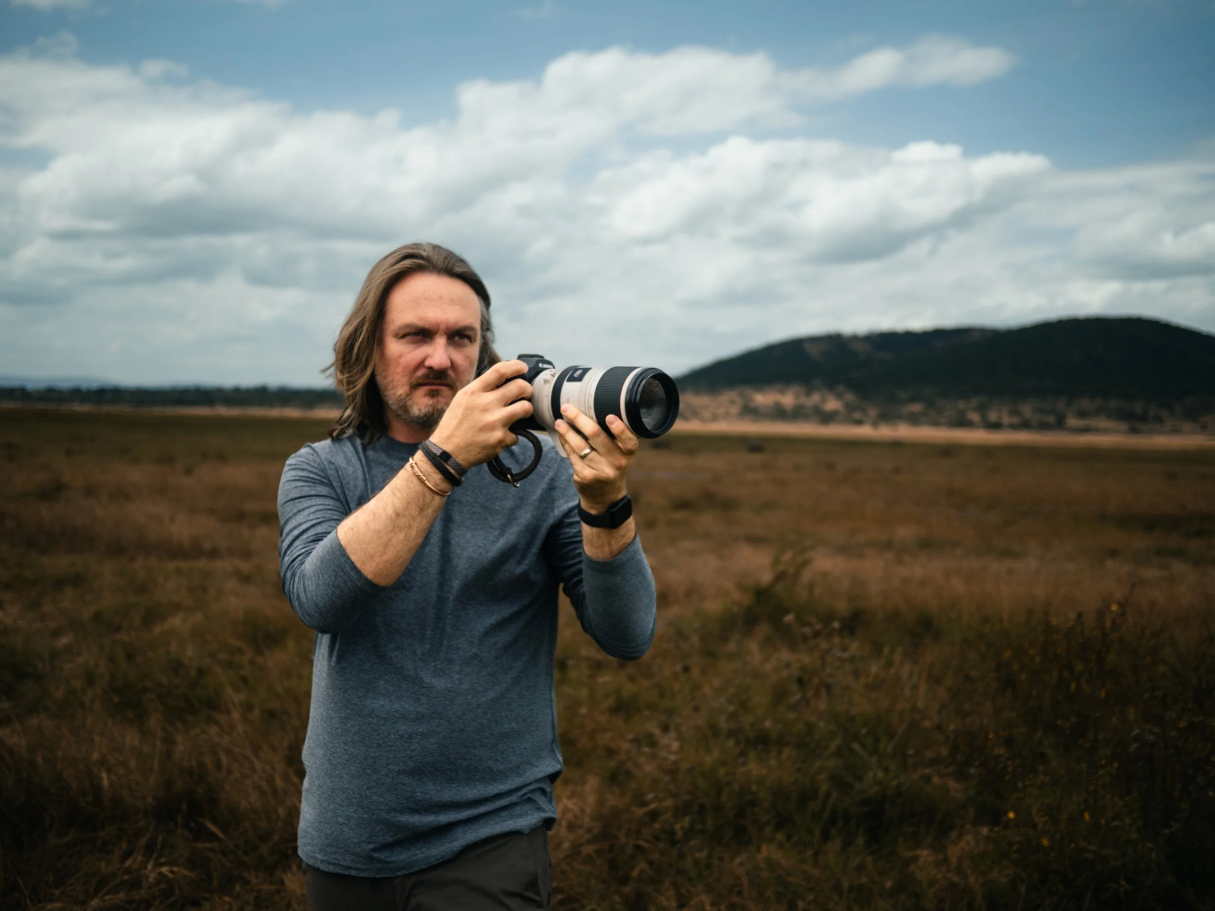 Photographer holding a telephoto camera in a grassy field with mountains under a cloudy sky