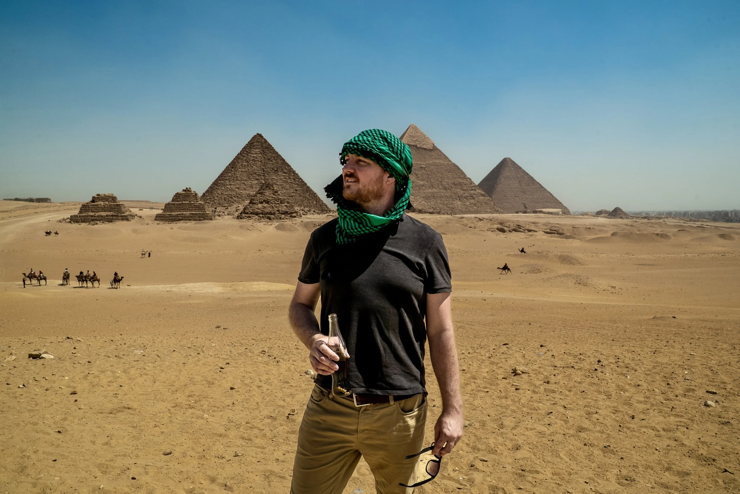 Traveler wearing a green headscarf stands in Egypt’s Giza desert holding a bottle, with the pyramids and camel riders behind him.