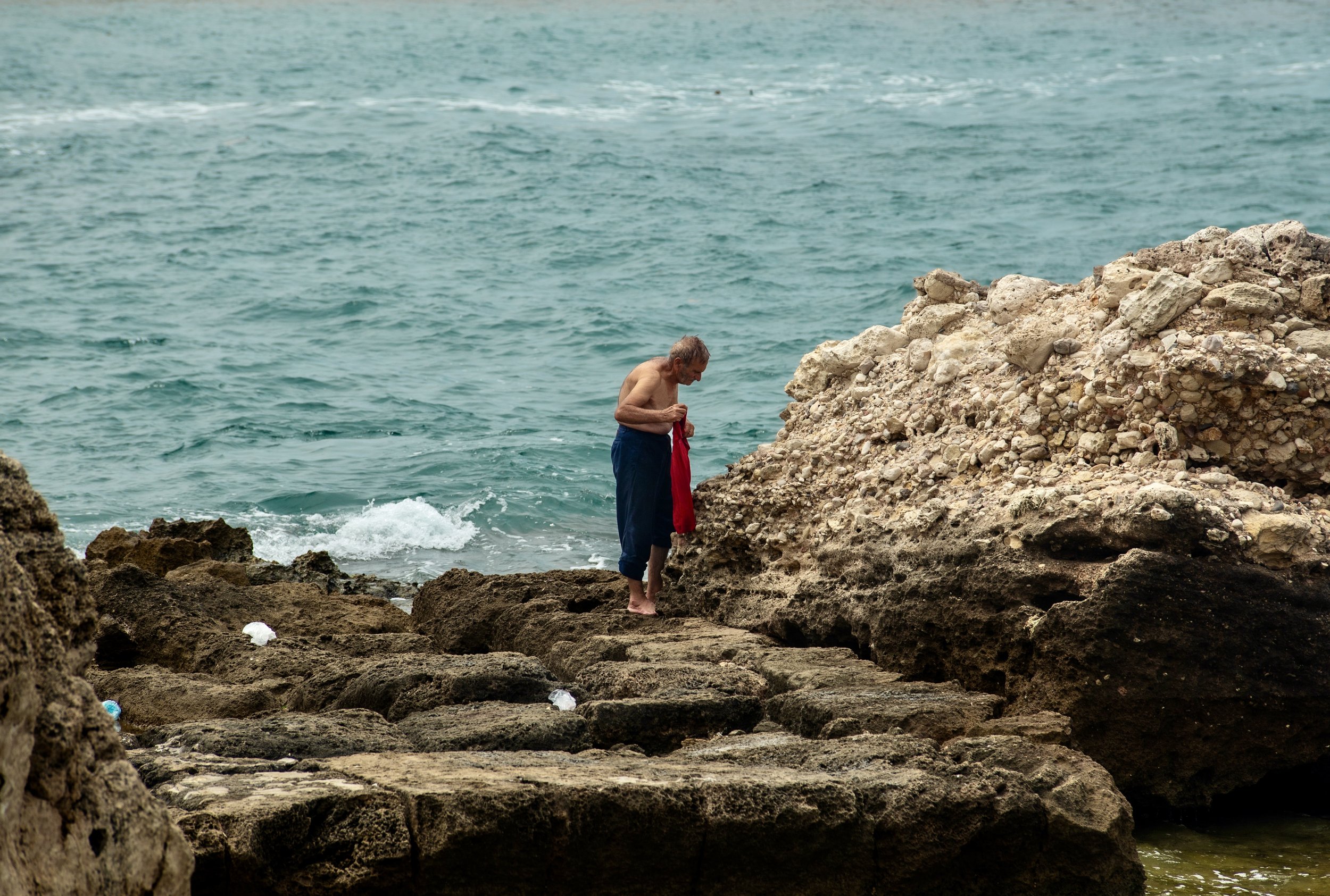Shirtless man holding a red towel on rocky seashore beside turquoise ocean waves in Lebanon. Photo by David Ferry Jr.