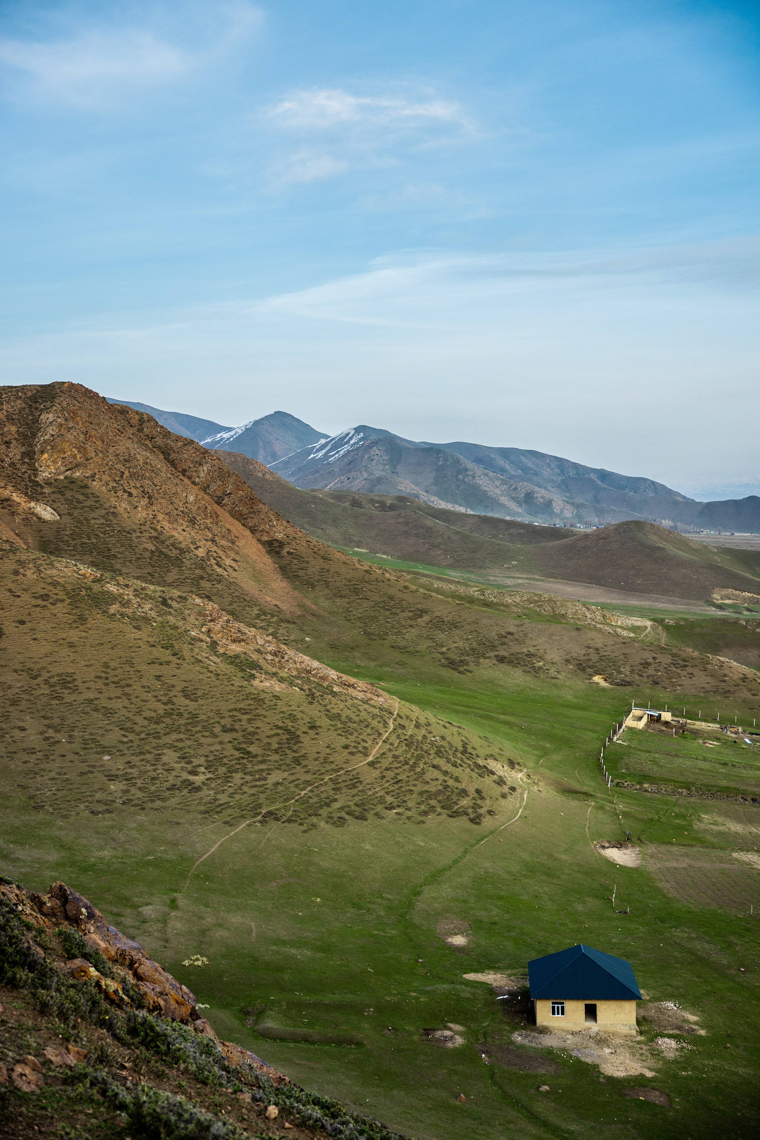 Mountain valley with snow-capped peaks, green rolling hills, dirt paths, and a small blue-roofed house under a bright blue sky in Kyrgyzstan. Photo by David Ferry Jr.
