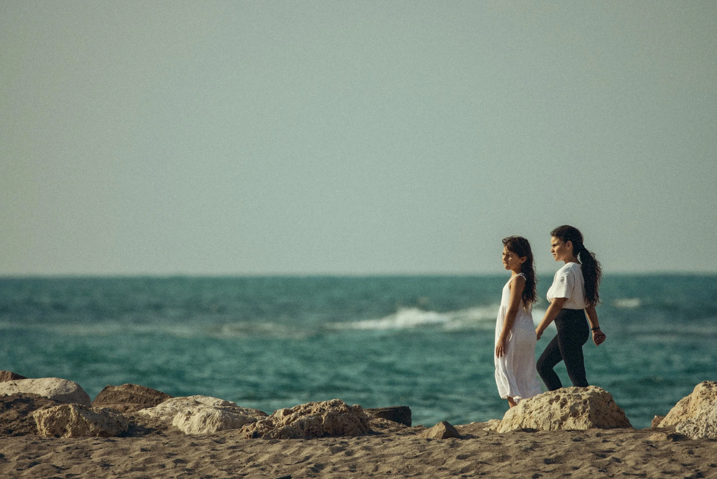Two children holding hands while walking along rocky seaside cliffs with ocean waves behind them in Israel. Photo by David Ferry Jr.