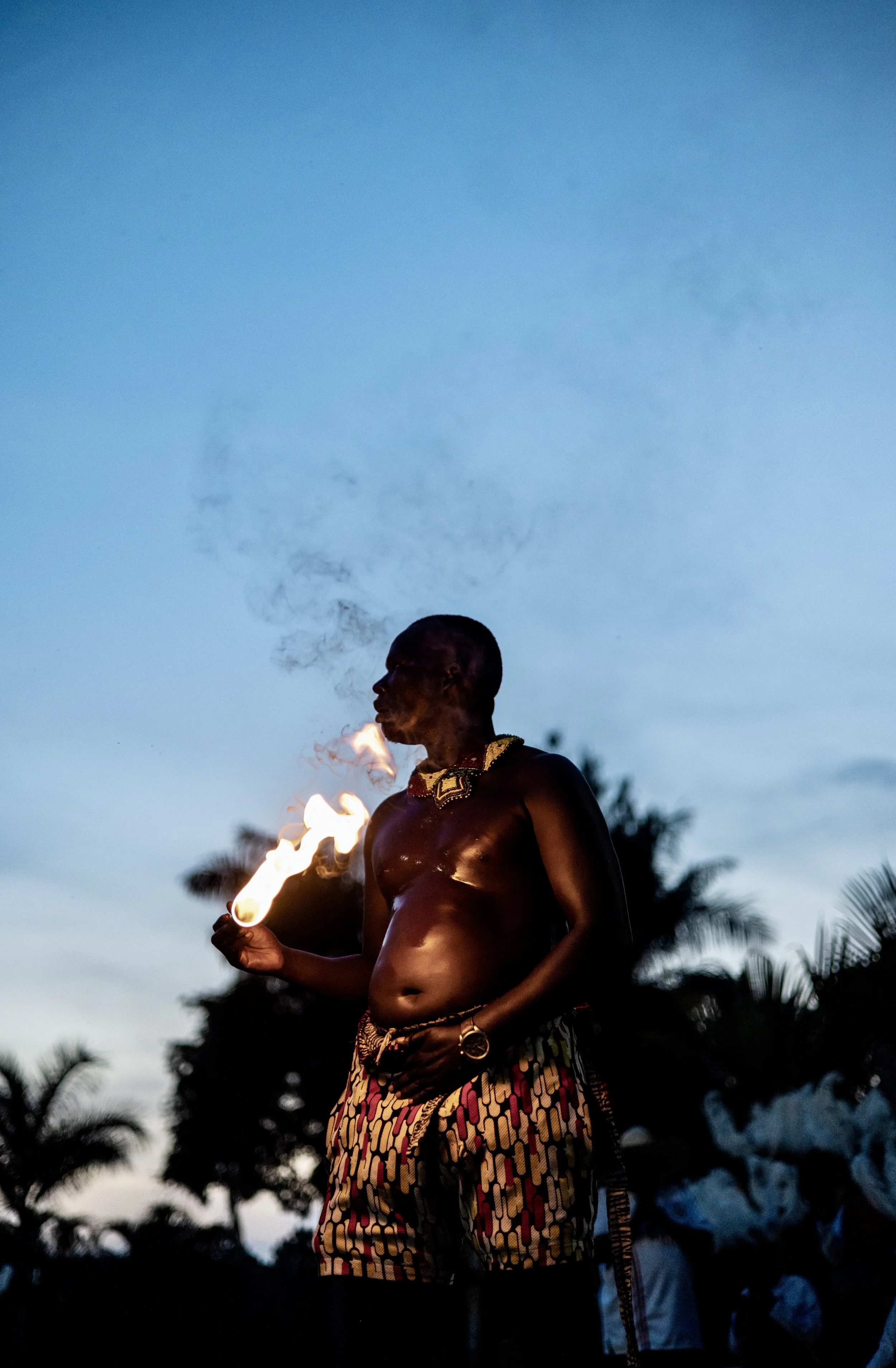 Shirtless man in patterned traditional clothing holding a flaming torch outdoors at dusk