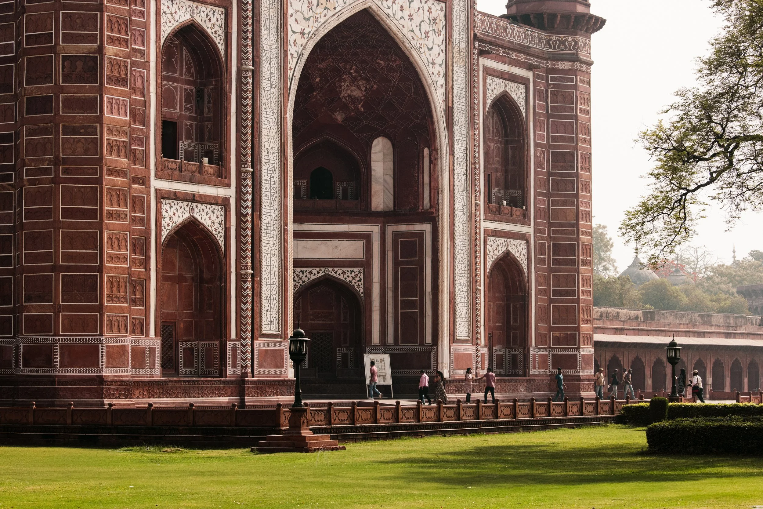 Ornate red sandstone Mughal architecture with arched entrances, white calligraphy, visitors, and manicured gardens at the Taj Mahal complex in Agra, India. Photo by David Ferry Jr.
