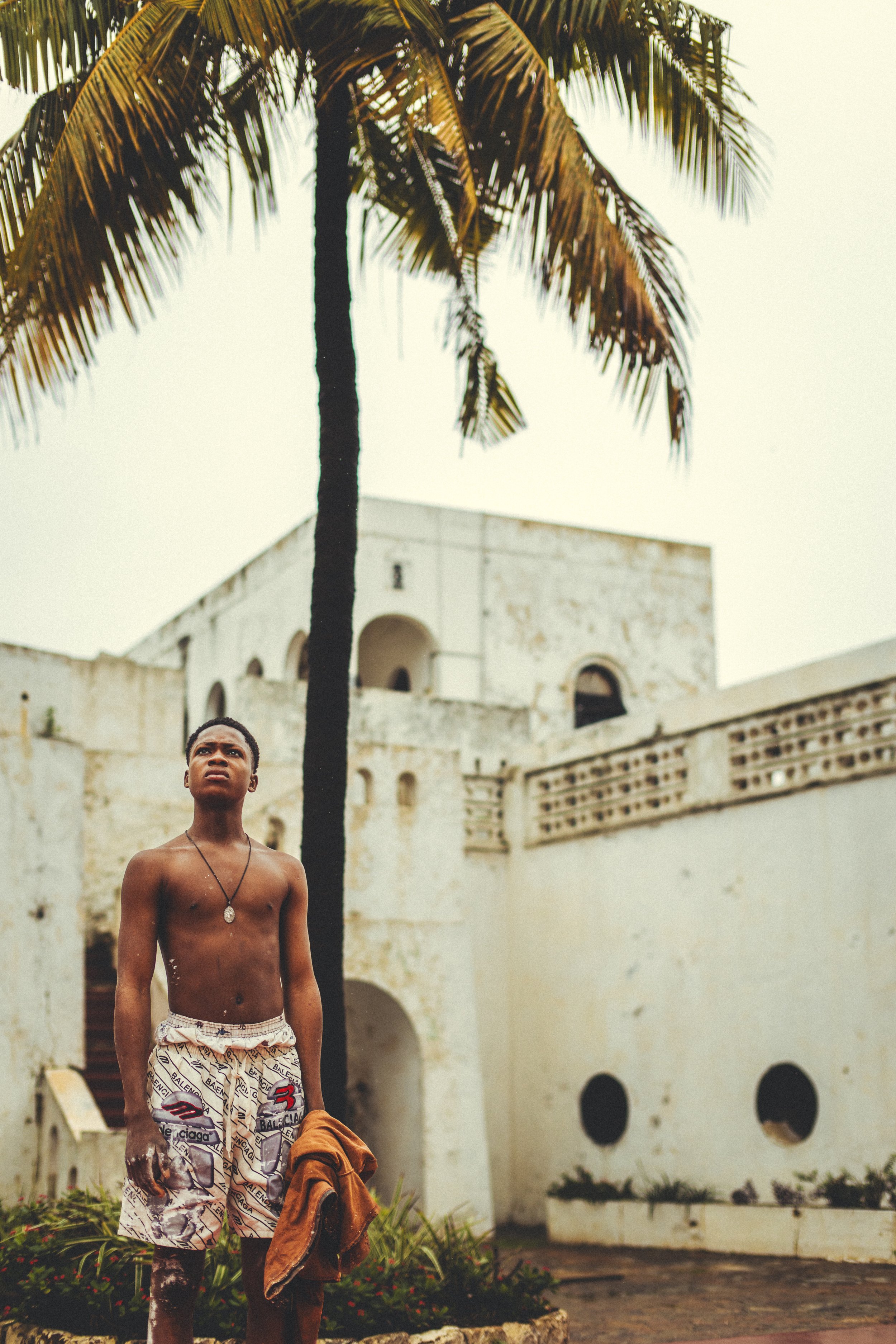 Shirtless young man standing beneath a palm tree in front of a weathered colonial building in Accra, Ghana. Photo by David Ferry Jr.