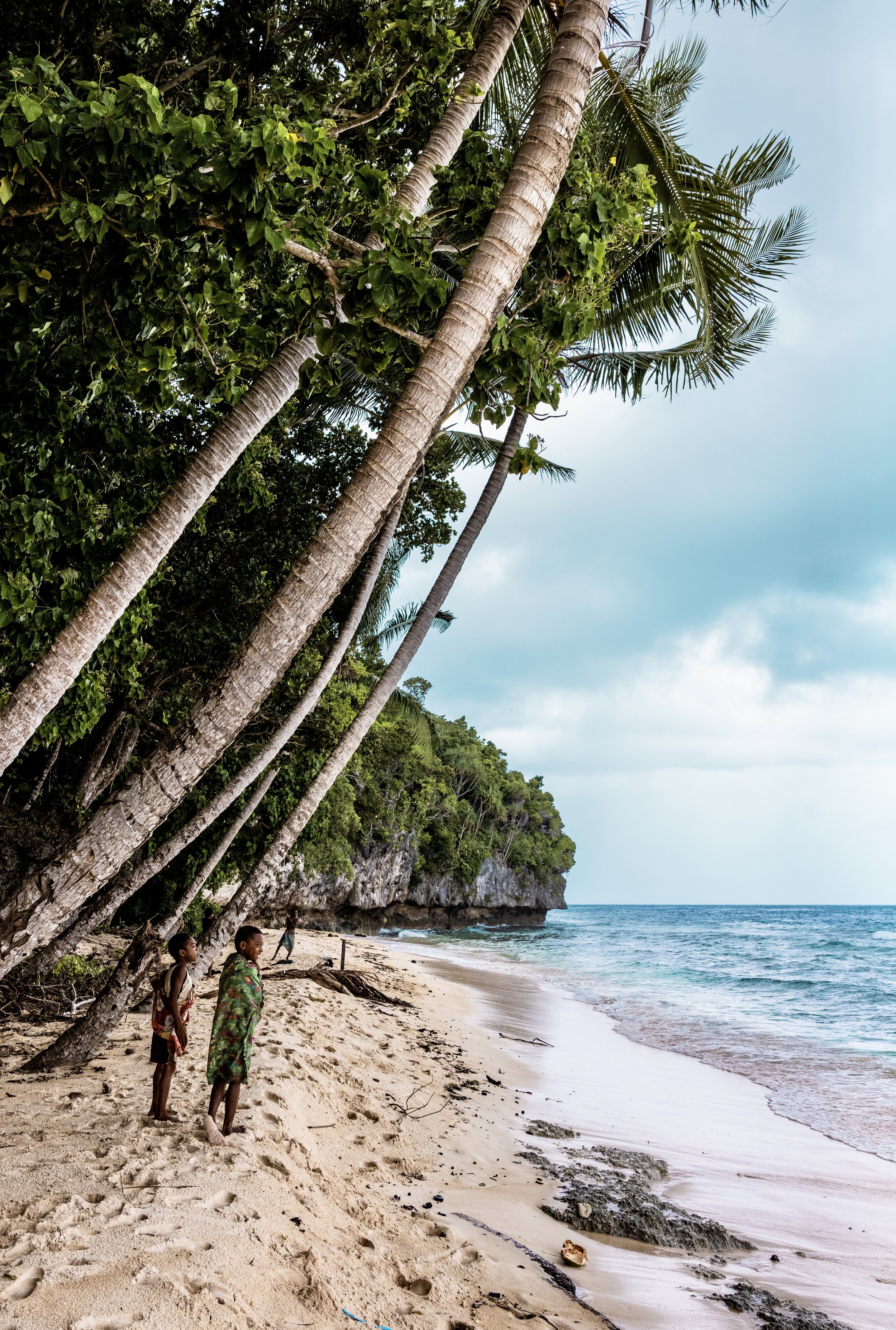 Tropical beach with leaning coconut palms, lush greenery, turquoise waves, limestone cliffs, and children on the sand in Papua New Guinea. Photo by David Ferry Jr.