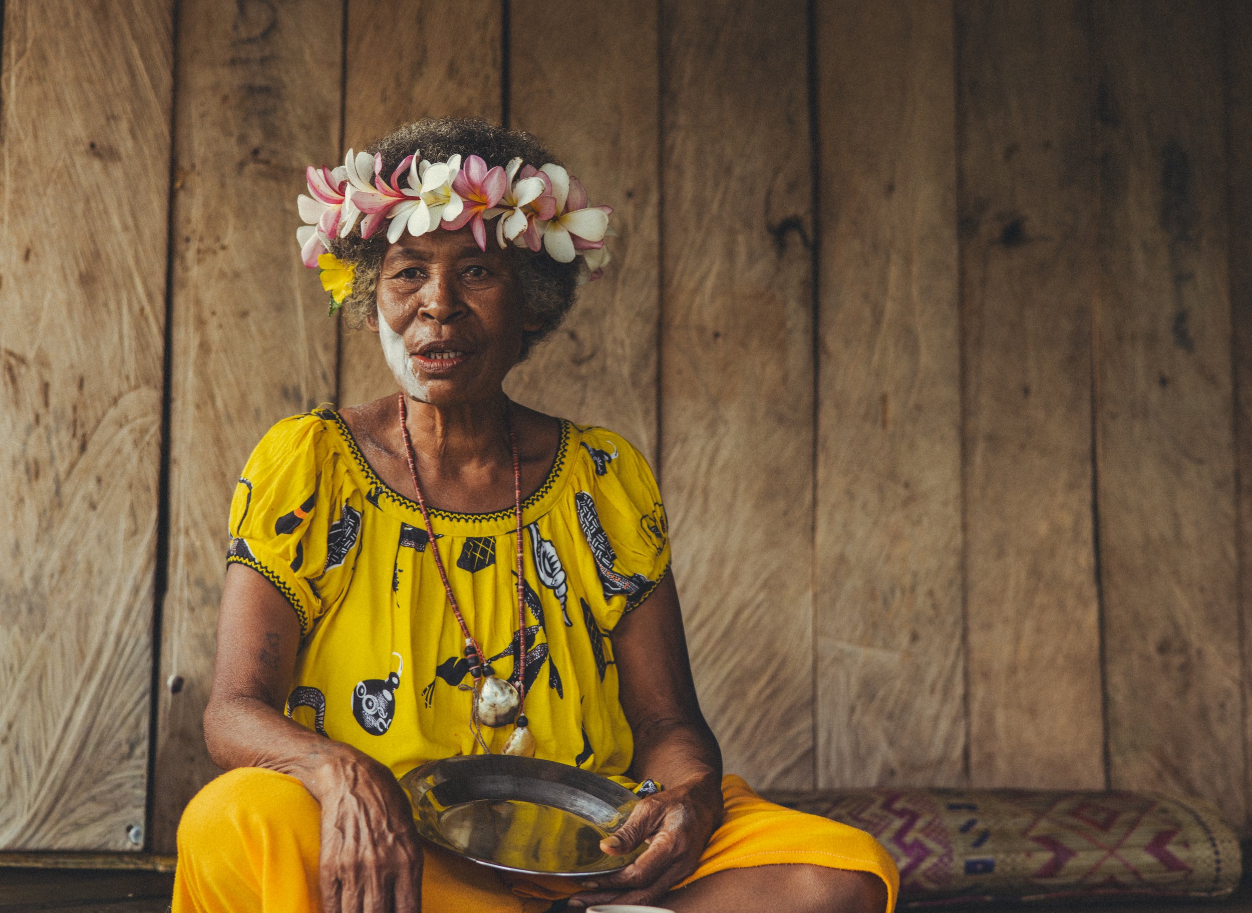 Indigenous woman wearing a flower crown and bright yellow traditional clothing, seated with a metal bowl inside a wooden home in Papua New Guinea. Photo by David Ferry Jr.