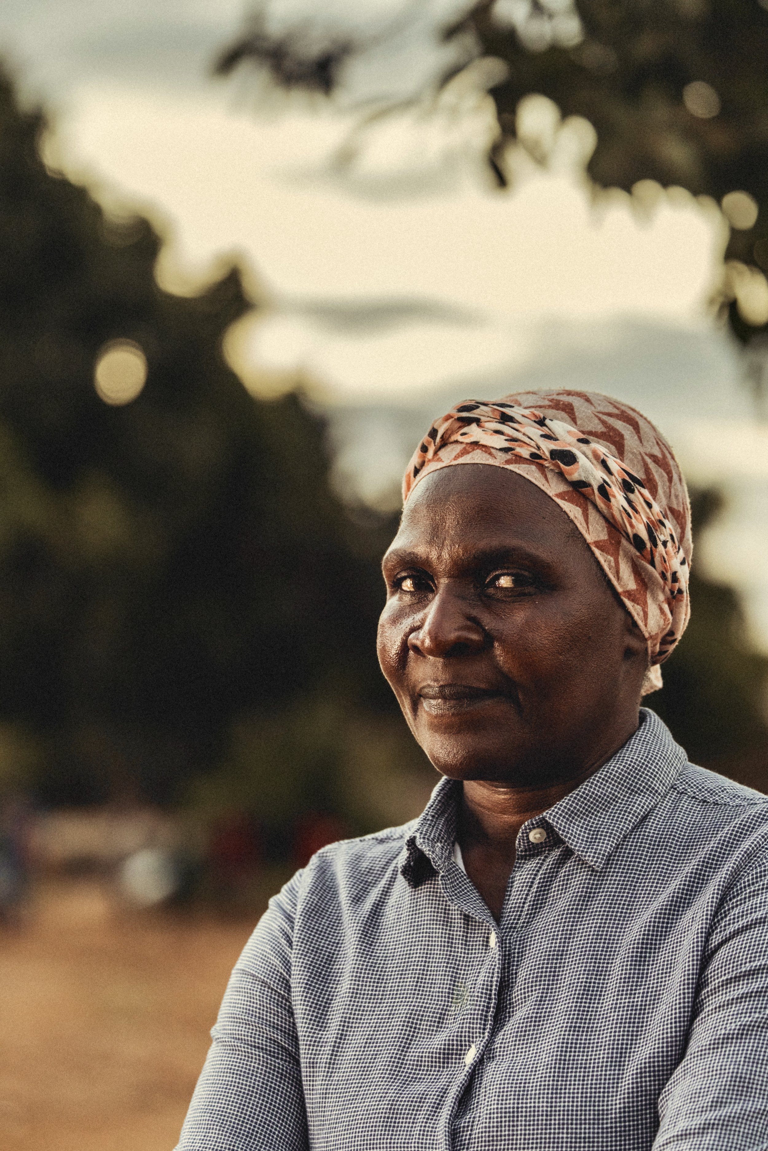 Portrait of an older woman from Zimbabwe wearing a patterned headscarf and checked shirt outdoors. Photo by David Ferry Jr.