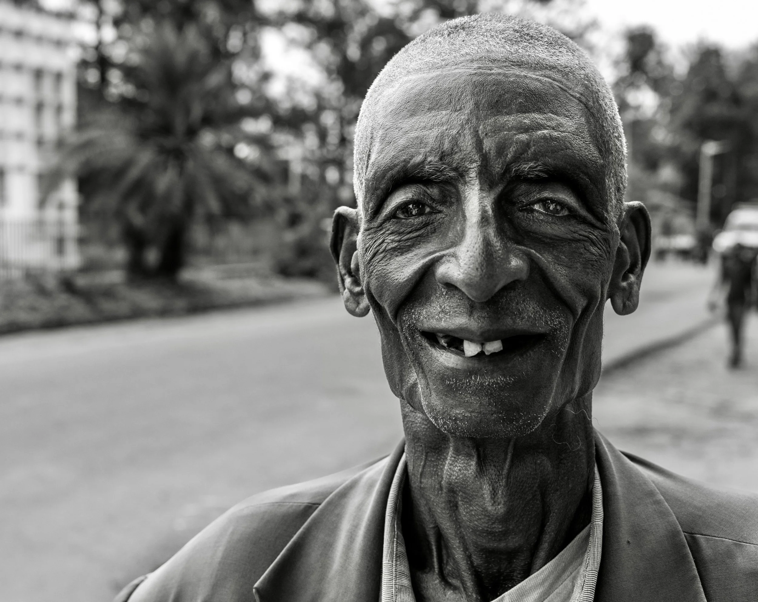 Black-and-white close-up portrait of a smiling older Ethiopian man outdoors, wearing a jacket and collared shirt. Photo by David Ferry Jr.