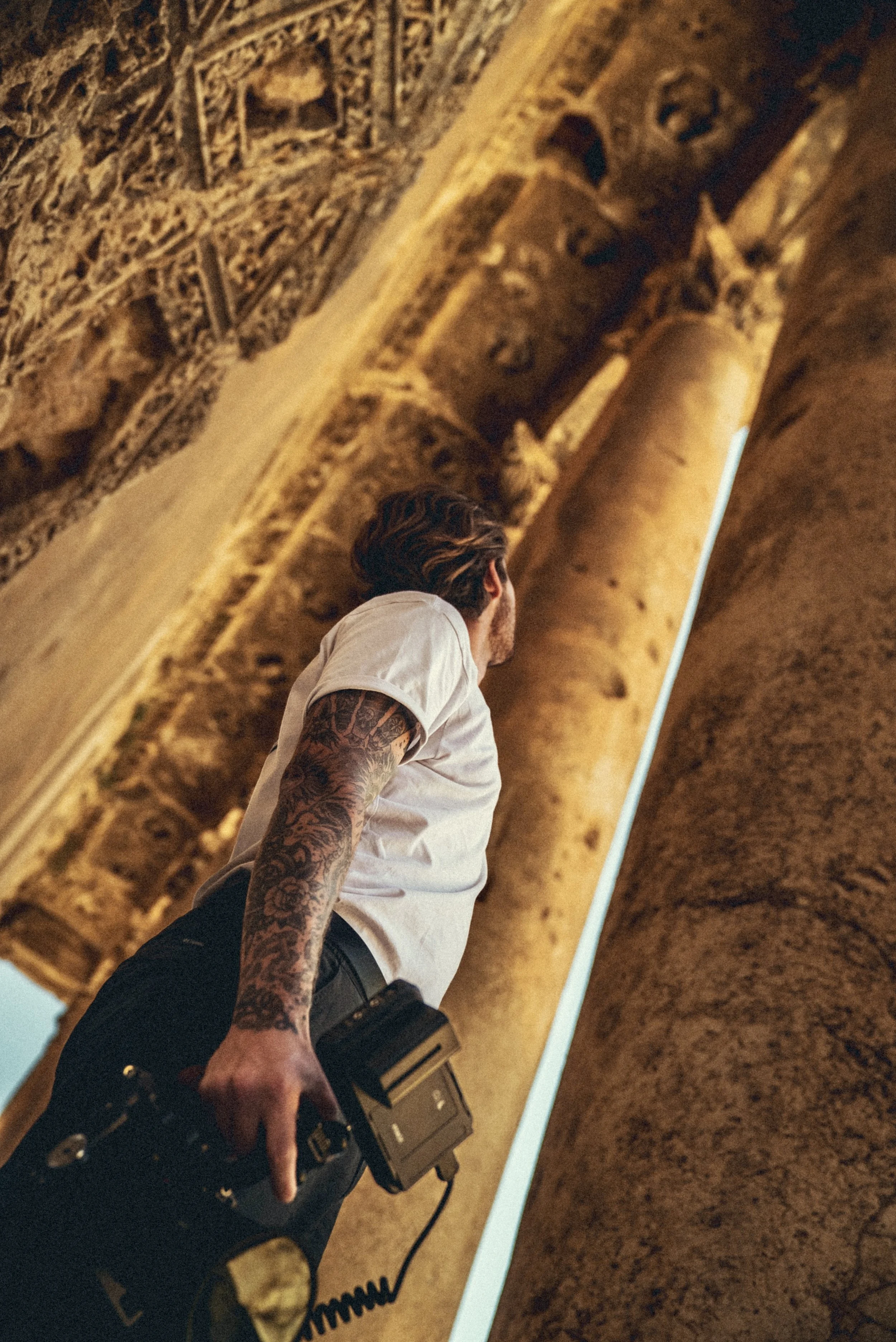 Tattooed man in a white shirt using a camera to photograph a large golden-brown mural or sculpture in Balbaak, Lebanon. Photo by David Ferry Jr.