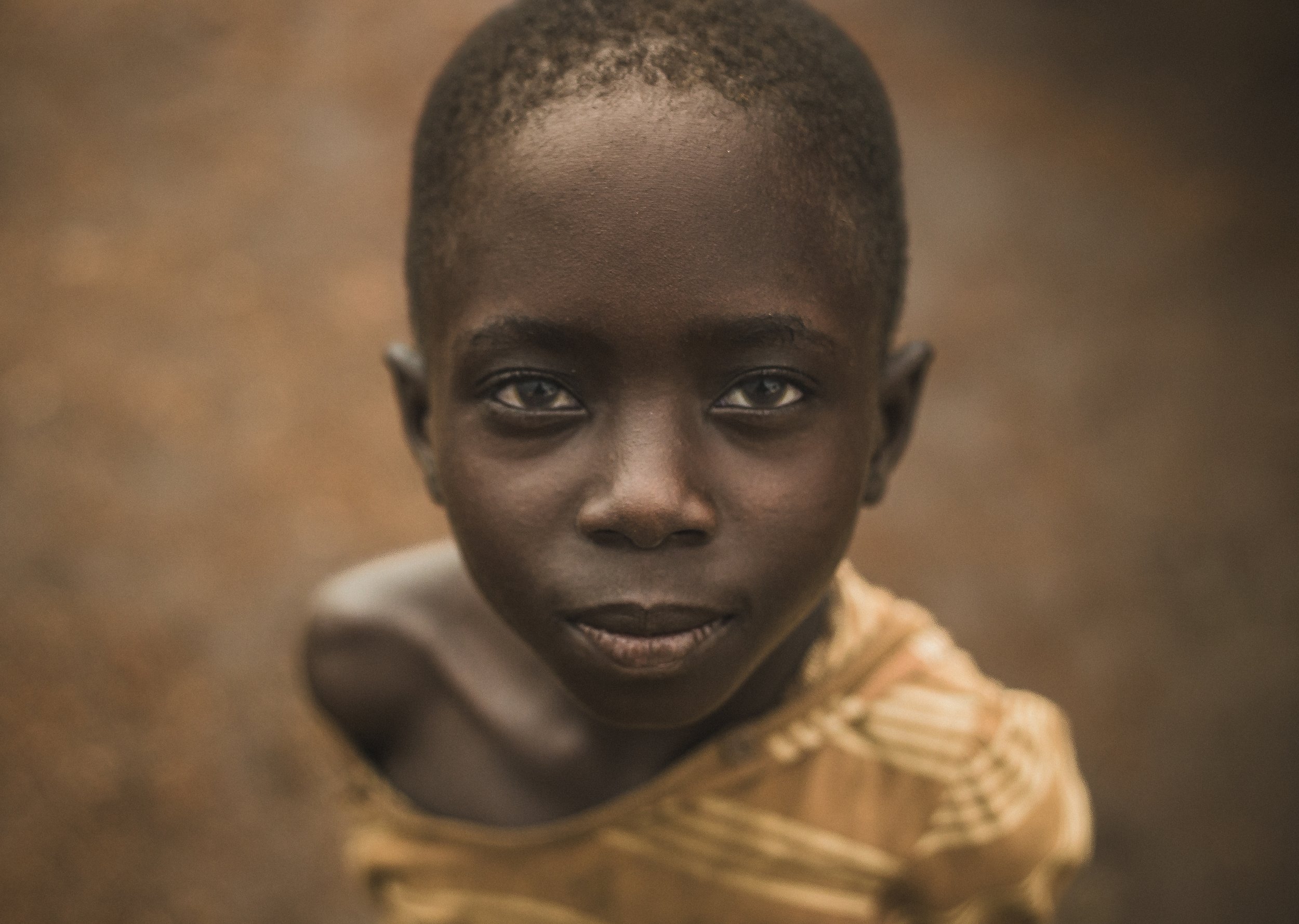 Close-up portrait of a smiling child with dark skin wearing a tan shirt in BidiBidi Refugee Settlement, Uganda. Photo by David Ferry Jr.