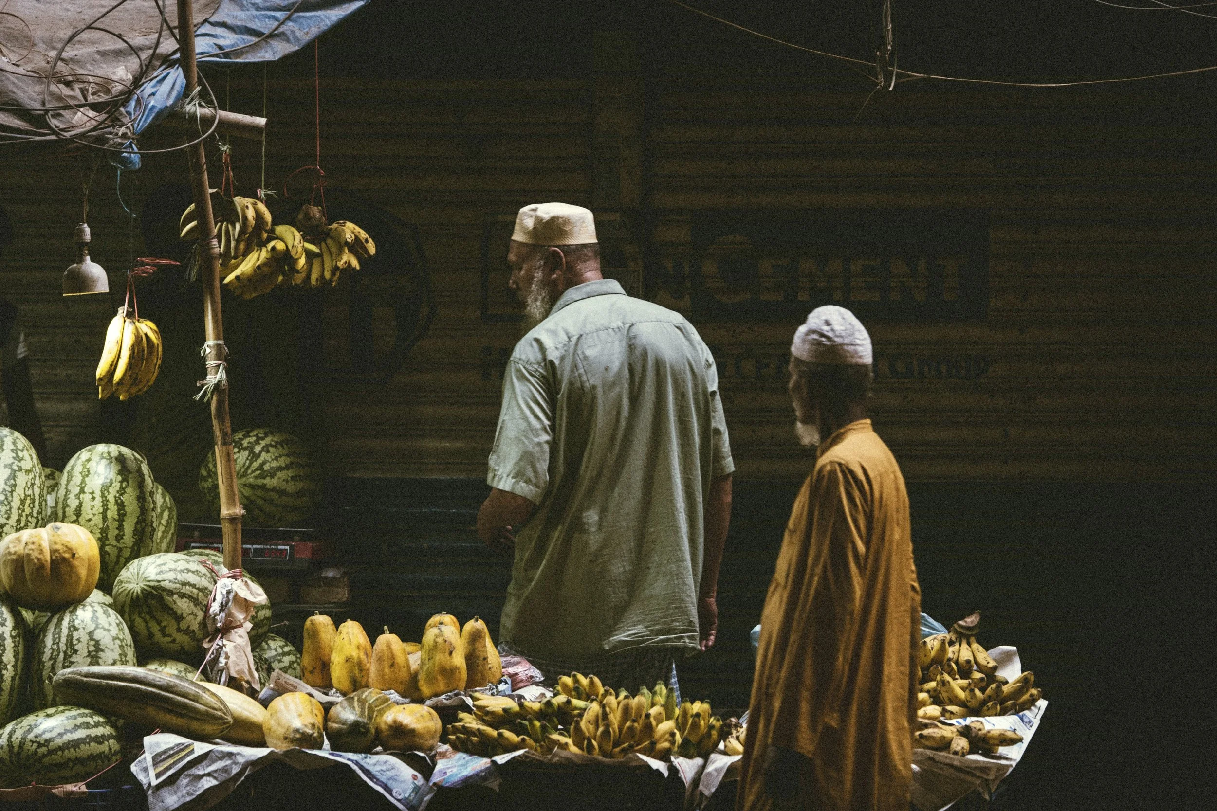 Two shoppers at a dimly lit market stall displaying bananas, watermelons, squash, and other fresh produce in Dhaka, Bangladesh. Photo by David Ferry Jr.