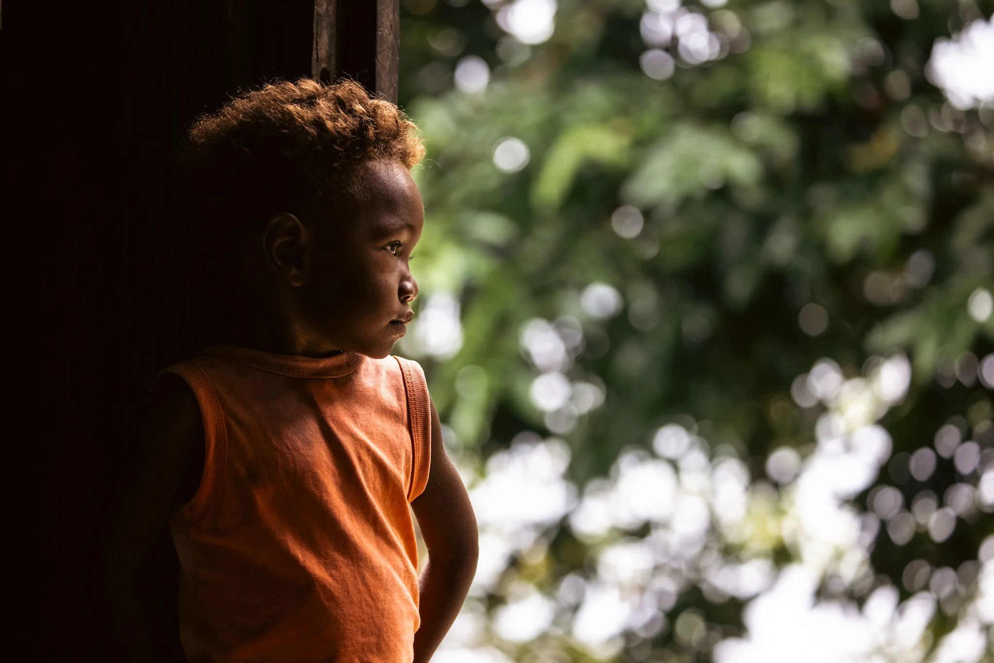 Young Black child in an orange sleeveless shirt looking outdoors against a blurred leafy background