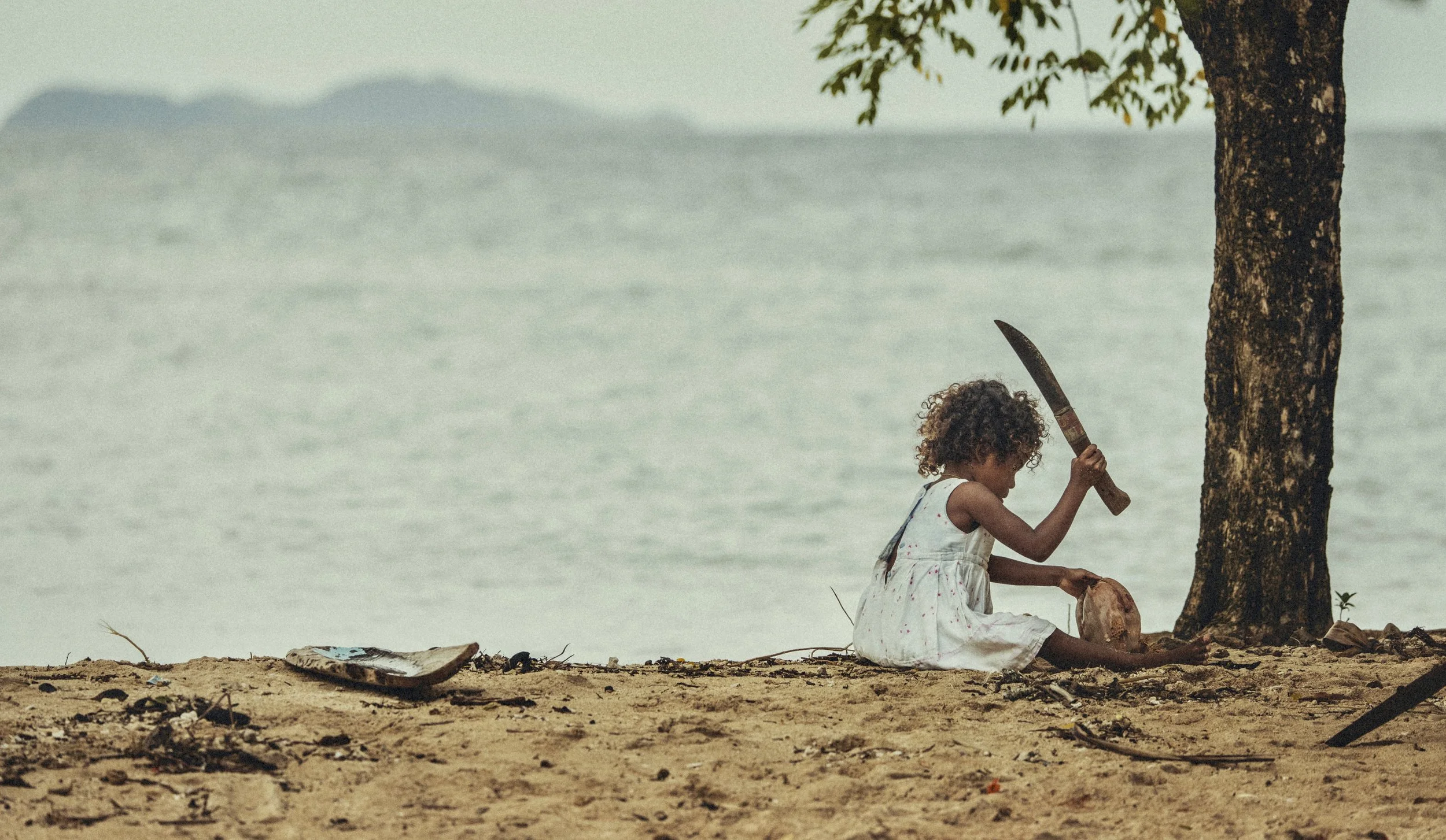 Child in a white dress playing with a coconut and machete beside a tree on a sandy beach in Papua New Guinea. Photo by David Ferry Jr.