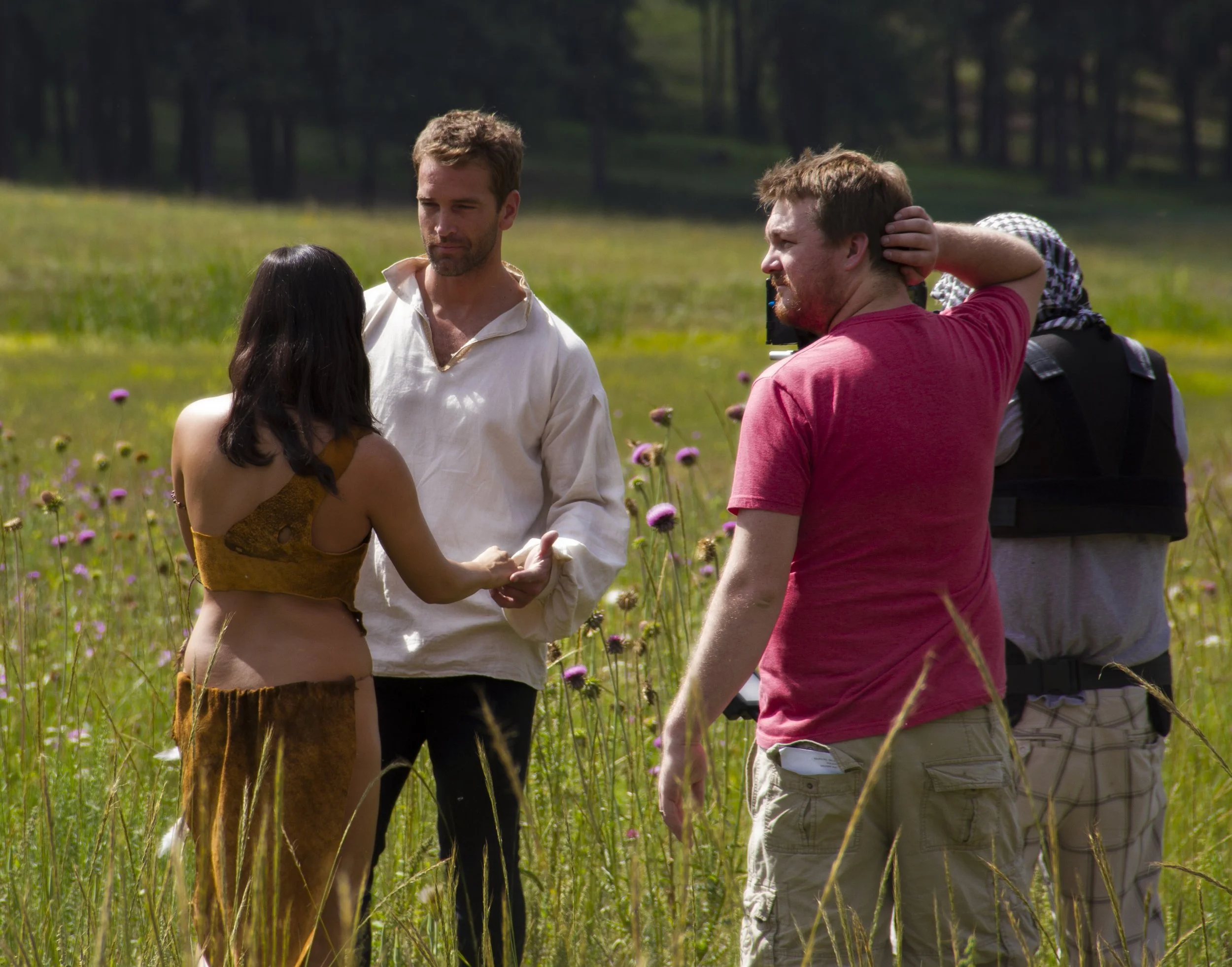Film crew filming actors in a wildflower meadow, with a camera operator adjusting a performer’s hair.