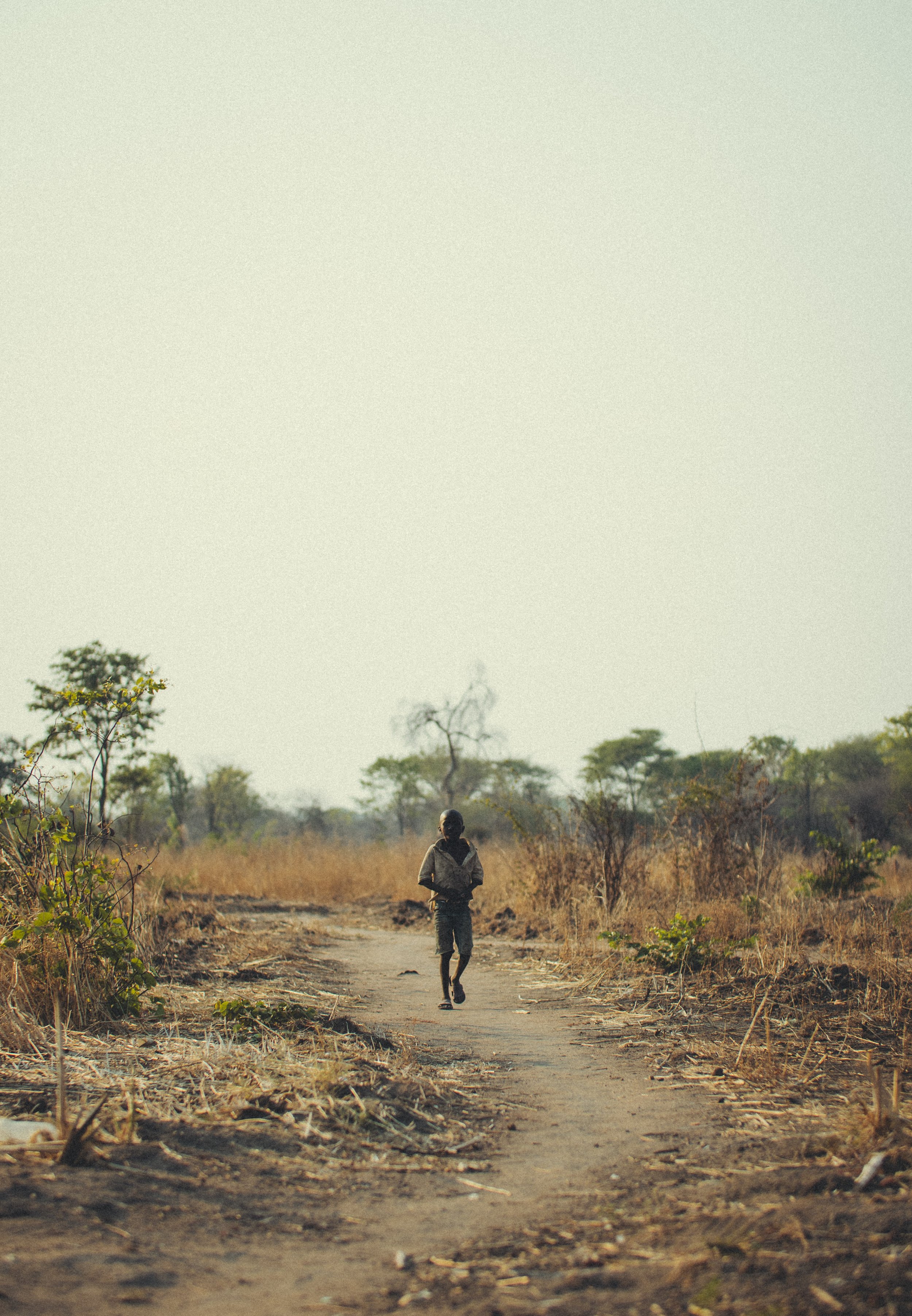 Child walking alone along a dirt path through a dry, wooded landscape in rural Zambia. Photo by David Ferry Jr.