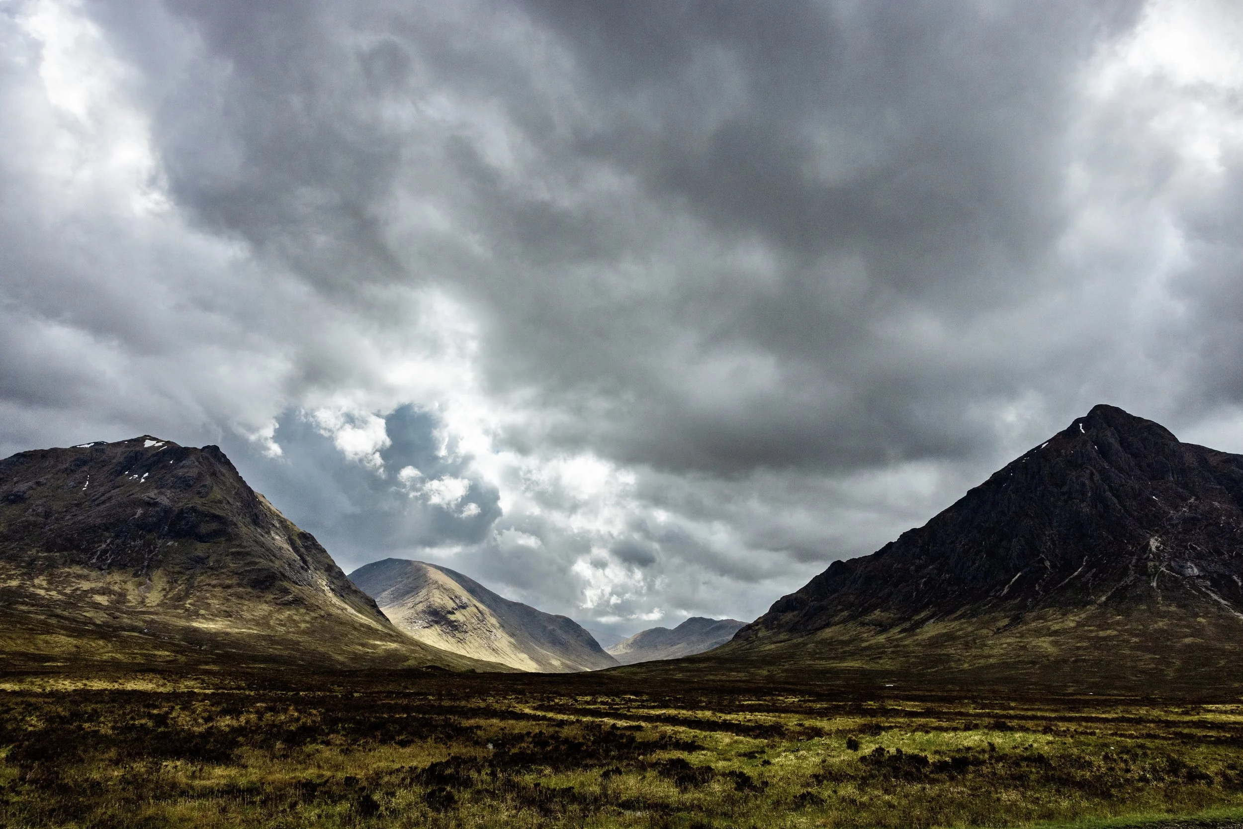 Dramatic cloudy mountain valley with rugged peaks, patches of snow, and green alpine landscape in Glencoe, Scotland. Photo by David Ferry Jr.