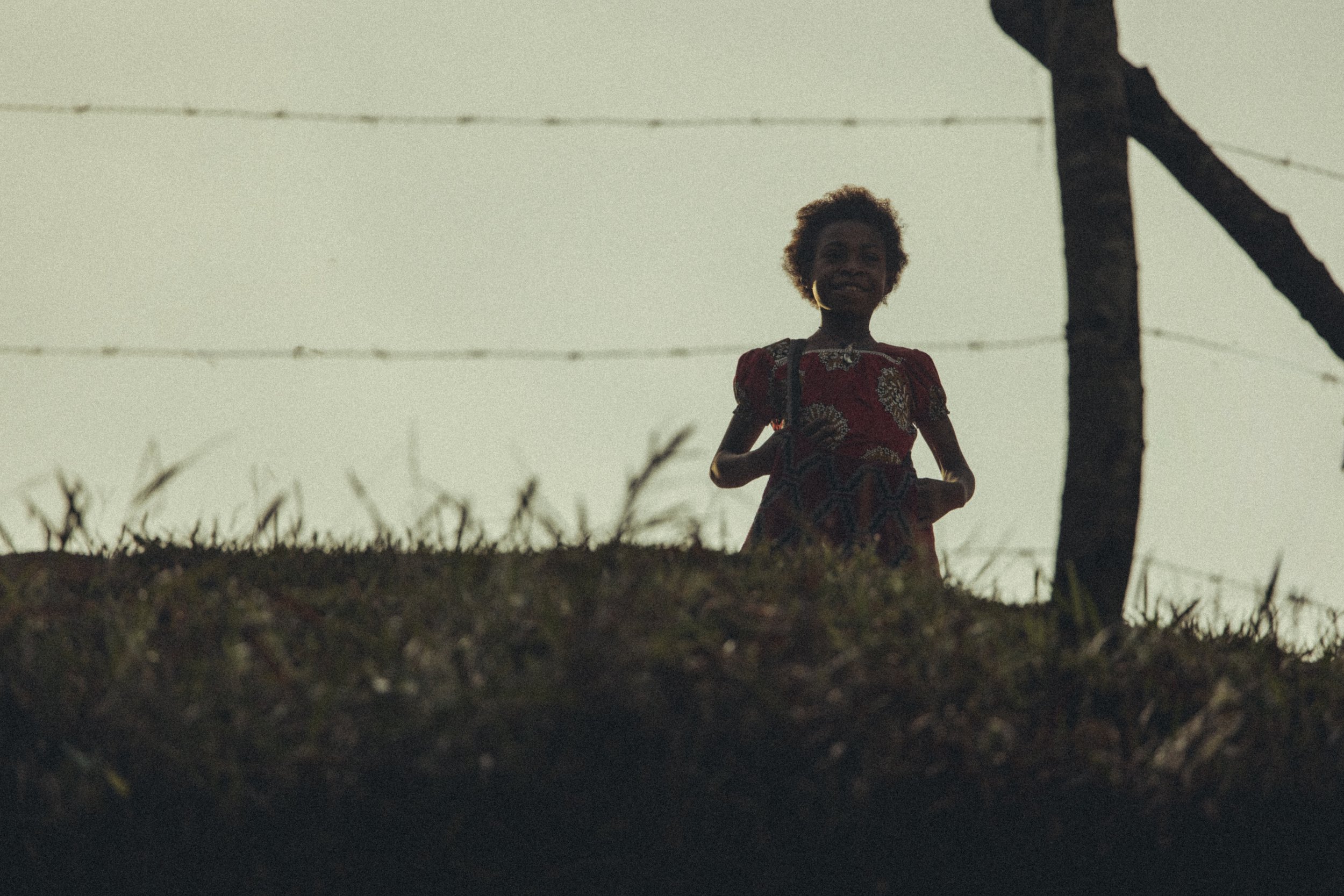 Low-angle portrait of a child in a red patterned dress standing in a grassy field beside a fence in the highlands of Papua New Guinea. Photo by David Ferry Jr.