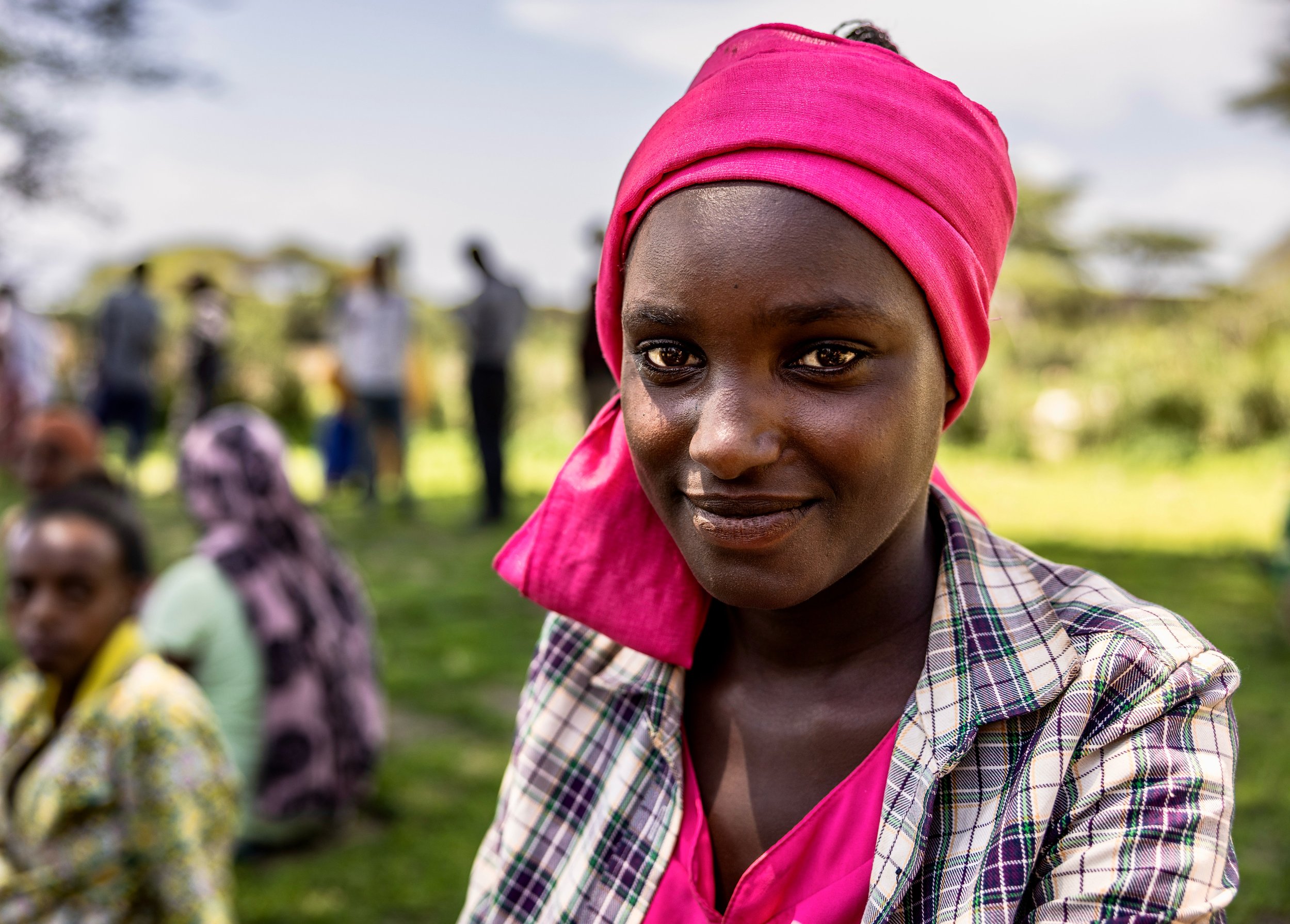 Close-up portrait of a smiling Black woman wearing a bright pink headscarf and plaid jacket outdoors, with people blurred in the background in Ethiopia. Photo by David Ferry Jr.