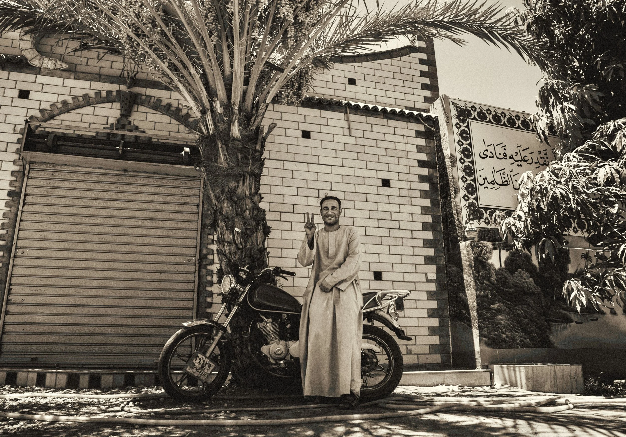 Sepia-toned photo of a smiling man in traditional clothing posing beside a motorcycle and date palm outside a brick house with Arabic writing in Egypt. Photo by David Ferry Jr.