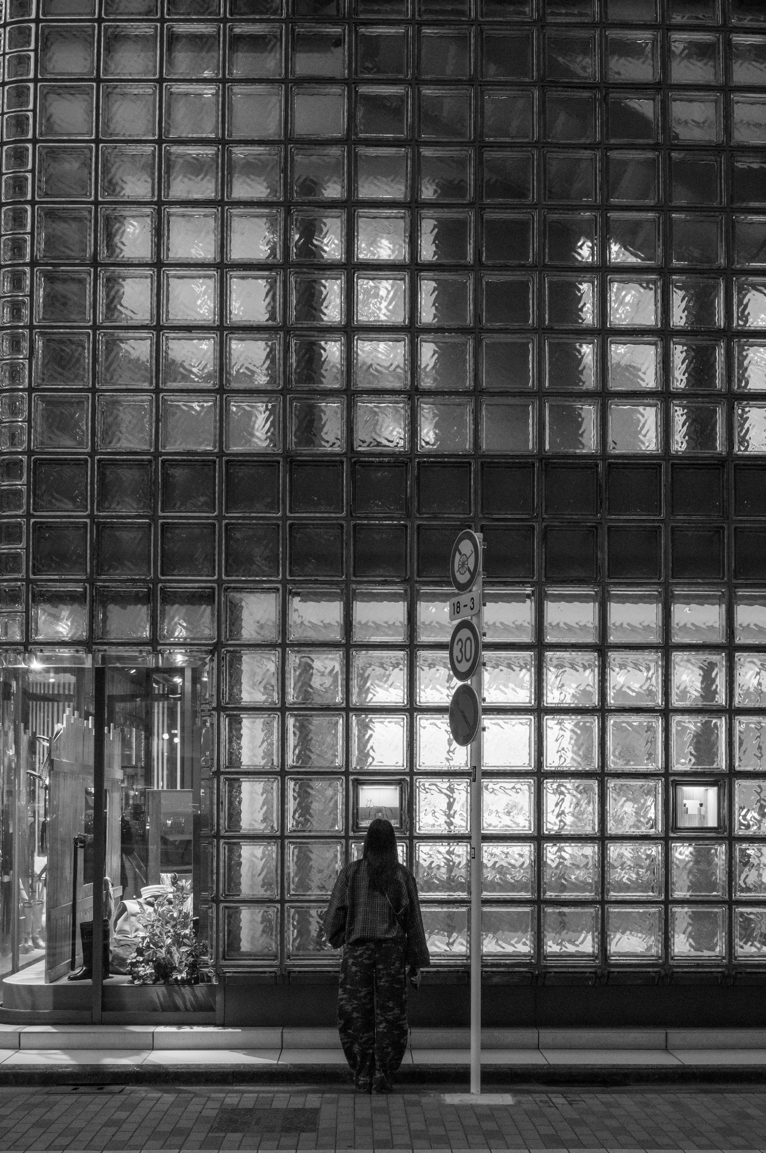 Black-and-white street scene with a person standing outside a storefront featuring a large glass-block wall, traffic signs, and a display window.