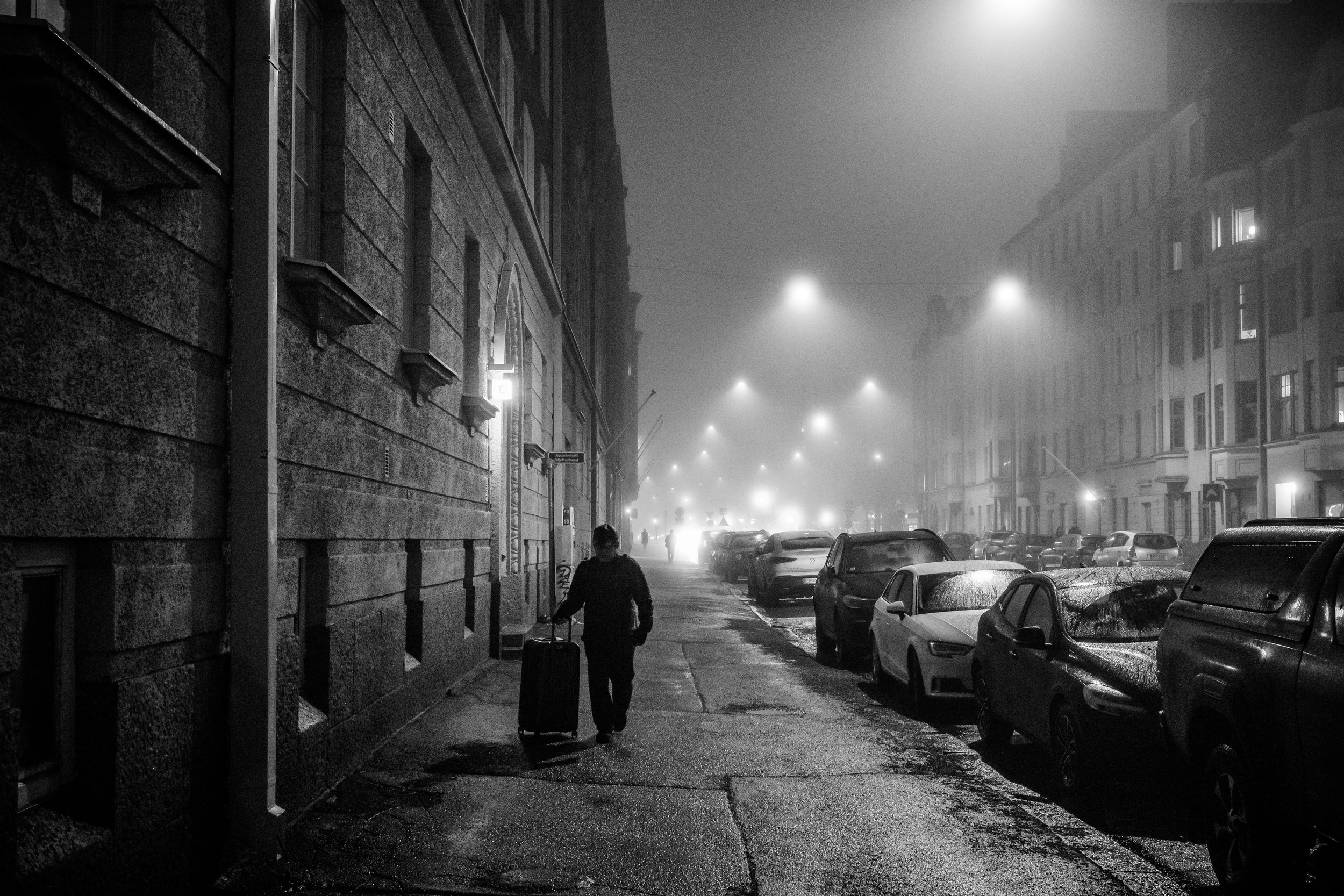 Black-and-white foggy city street at night with a traveler pulling a suitcase past parked cars and illuminated buildings
