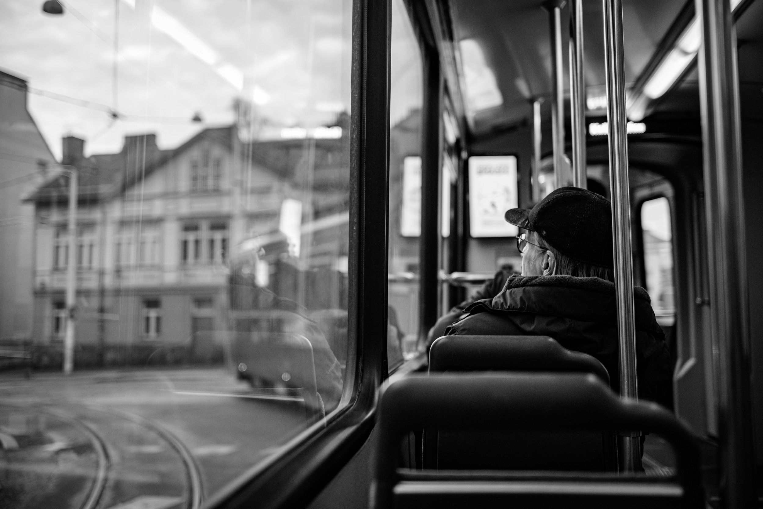Black-and-white view of an elderly passenger riding a city tram past blurred urban buildings