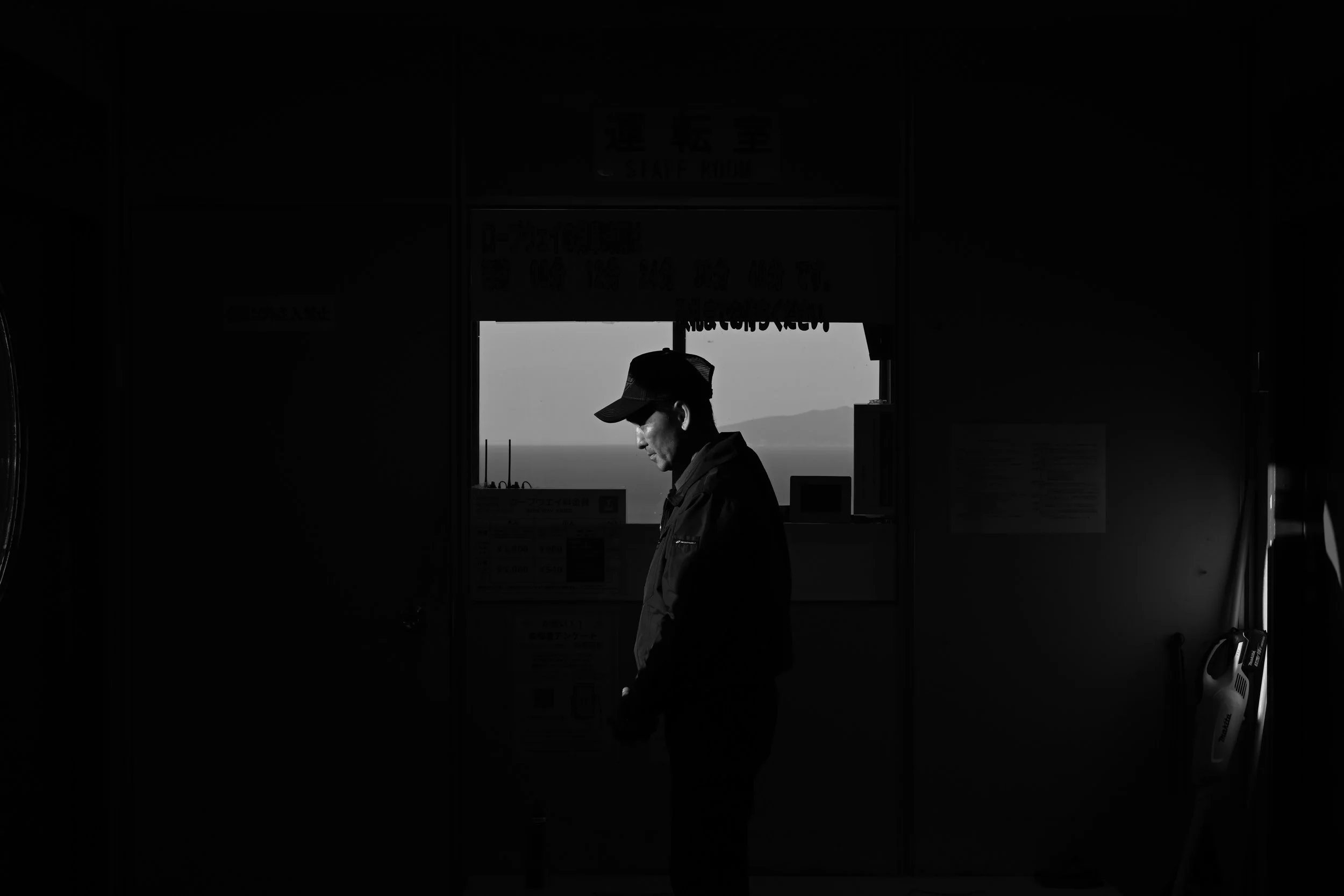 Black-and-white photo of a man in a baseball cap and jacket standing inside a dark building, with a mountain landscape visible through the doorway.