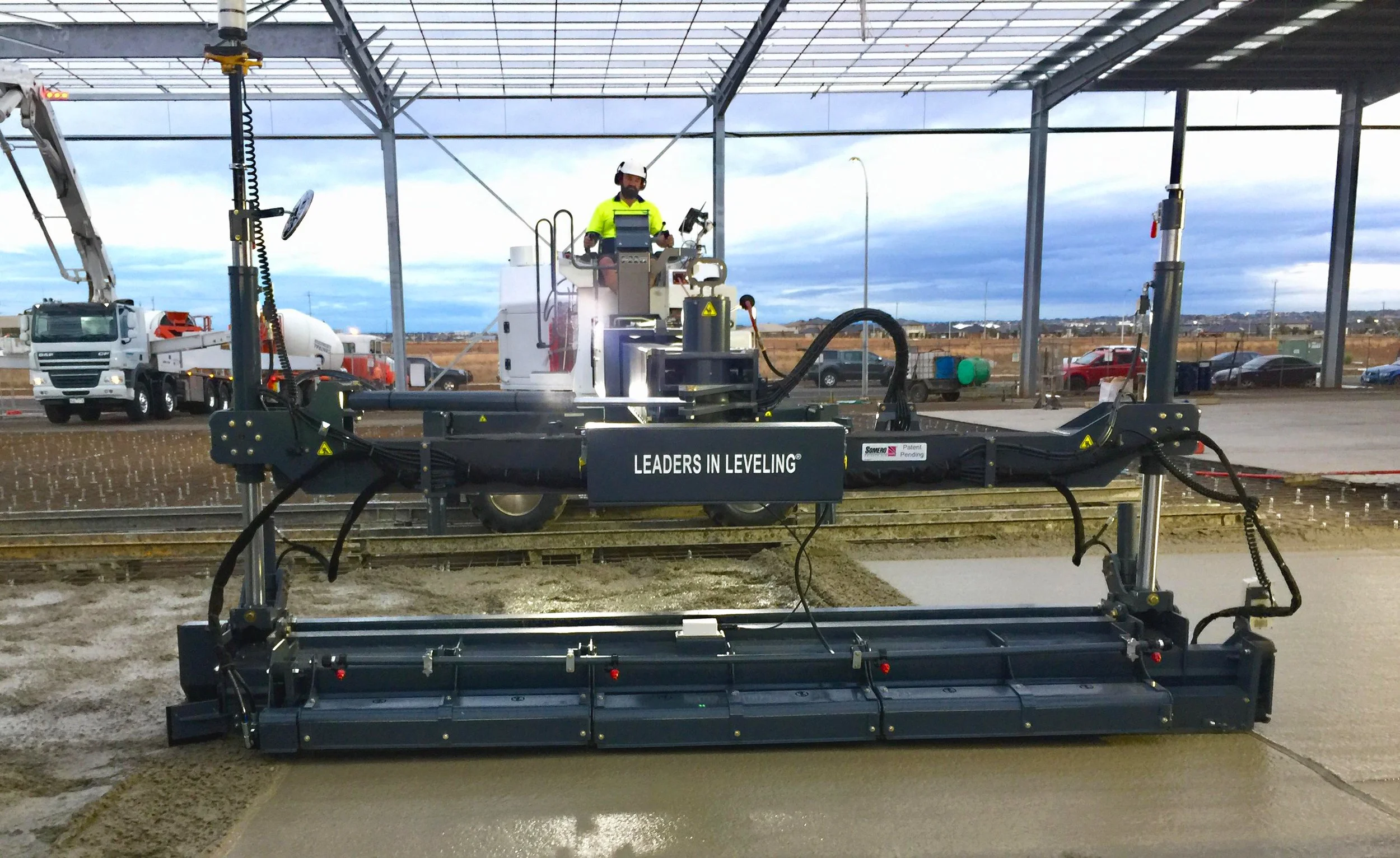 Concrete leveling machine at construction site, operated by a worker, with trucks in the background.