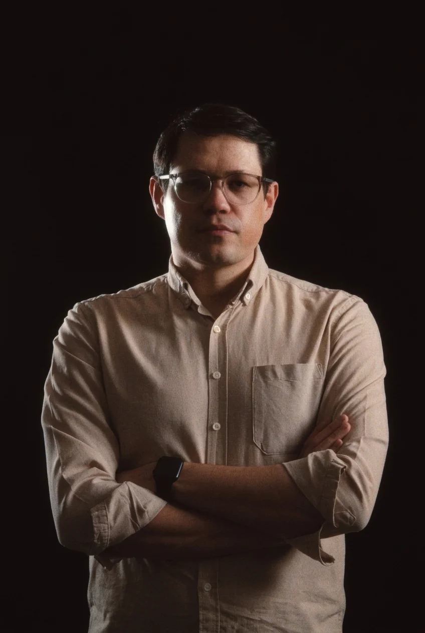 Portrait of a man wearing glasses and a beige button-down shirt with his arms crossed against a dark background