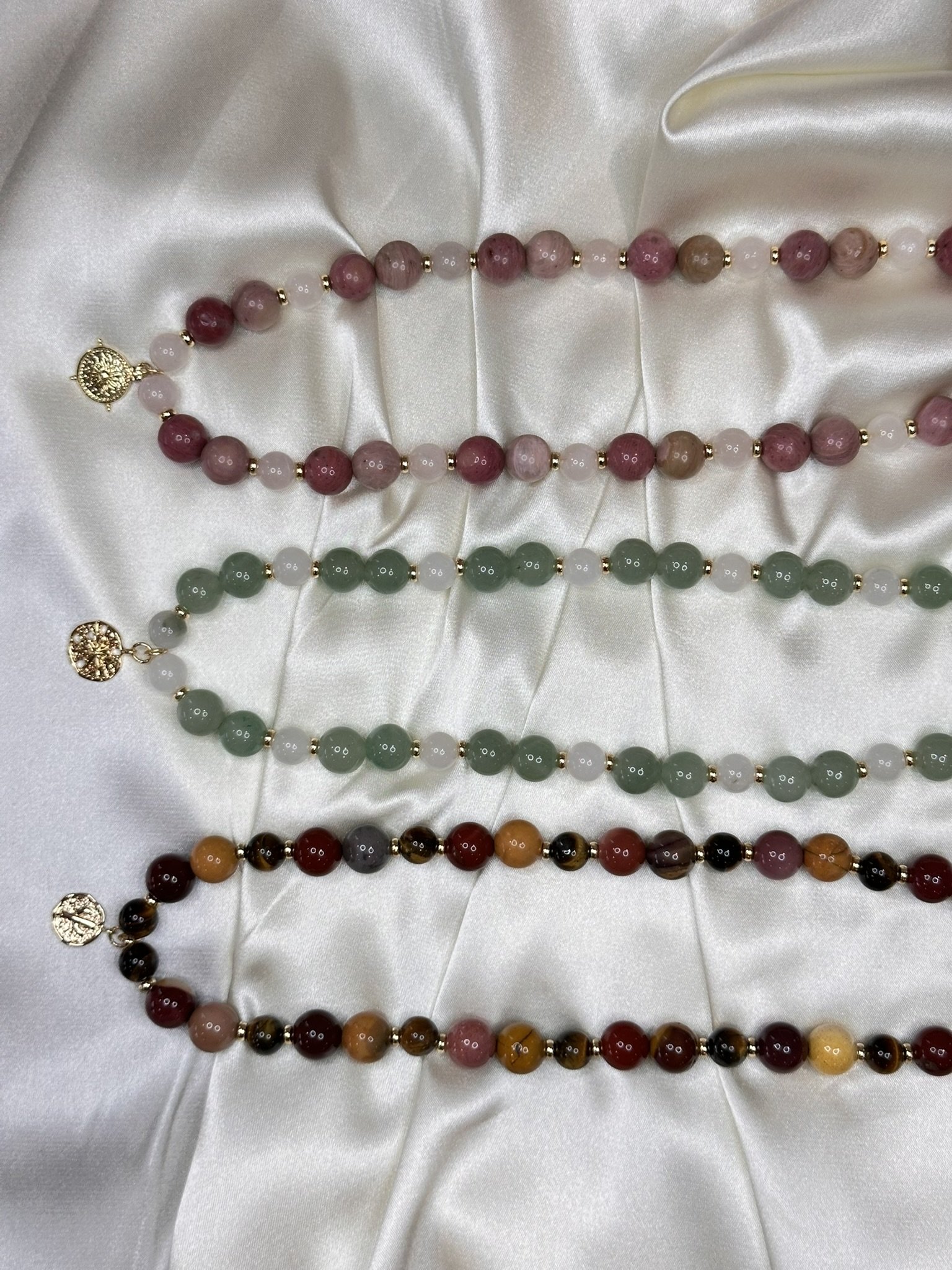 Three handmade gemstone beaded necklaces from Terra & Tide Collective, featuring genuine Amazonite, Tianshan Jade, Rhodonite, Rose Quartz, Mookaite and Tiger’s Eye gemstones with 14k gold-plated accents and charms, arranged on ivory satin.