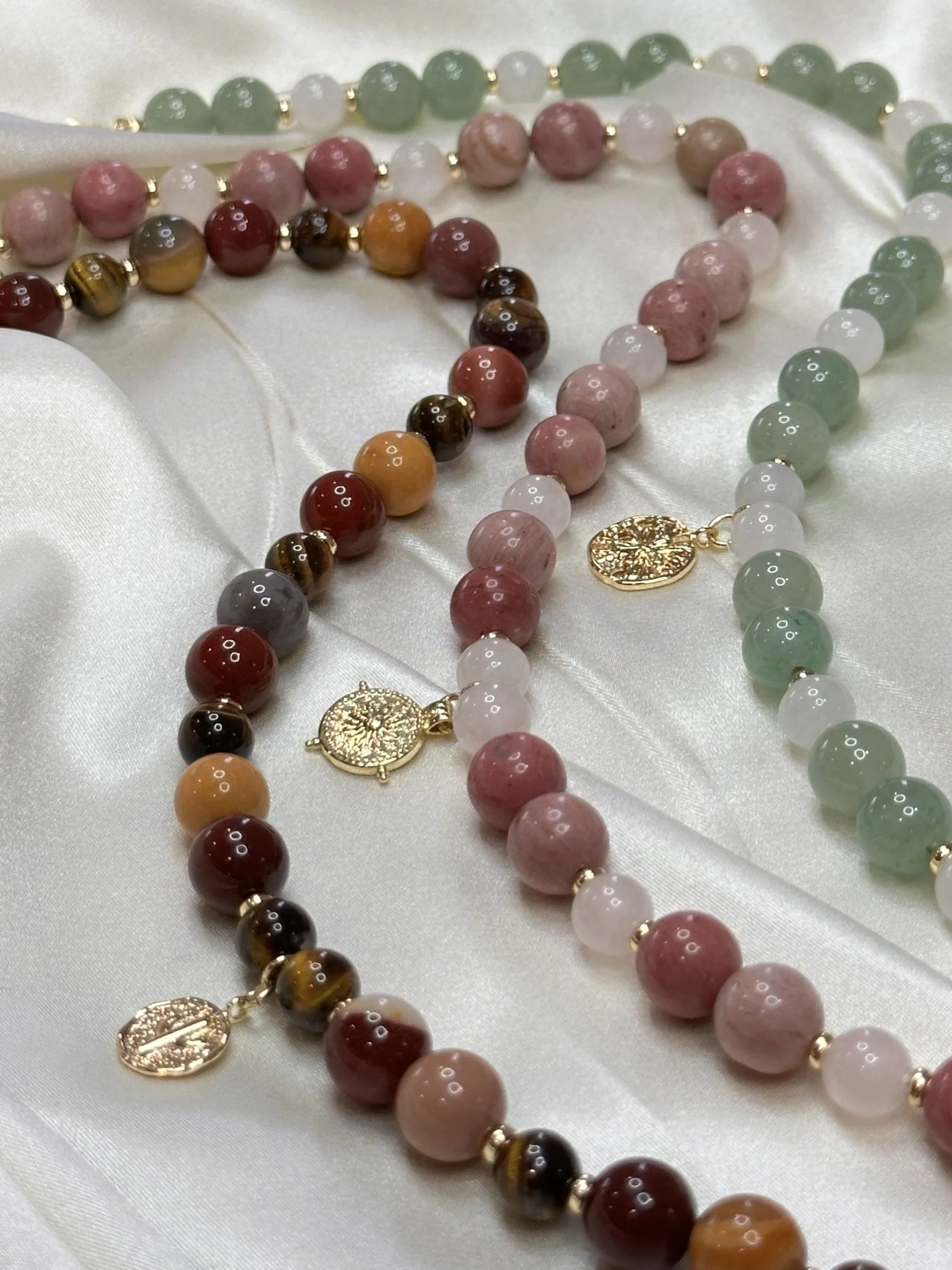 Three handmade genuine gemstone necklaces by Terra & Tide Collective, featuring Amazonite, Tianshan Jade, Rhodonite, Rose Quartz, Mookaite and Tiger’s Eye, with 14k gold-plated accents and charms, displayed on ivory satin.