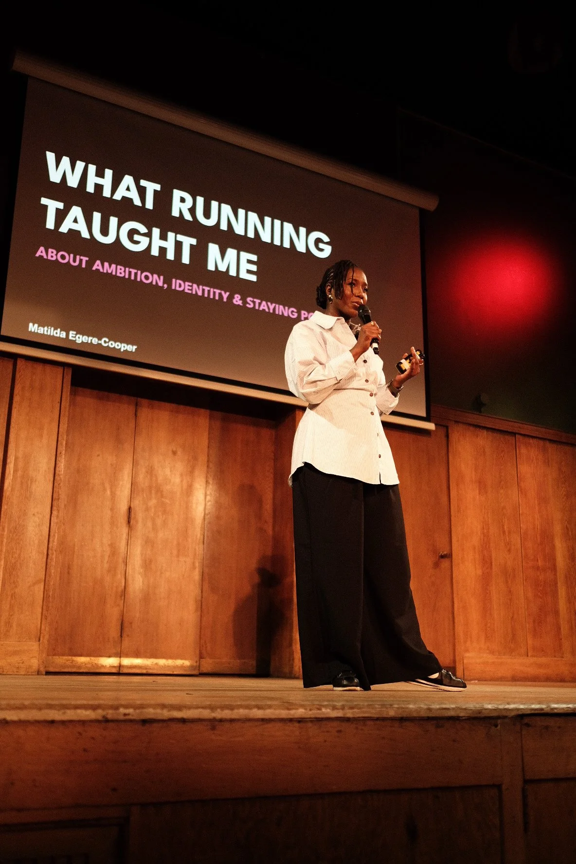 Speaker Matilda Egere-Cooper presents “What Running Taught Me” onstage, holding a microphone beneath a projected presentation slide.