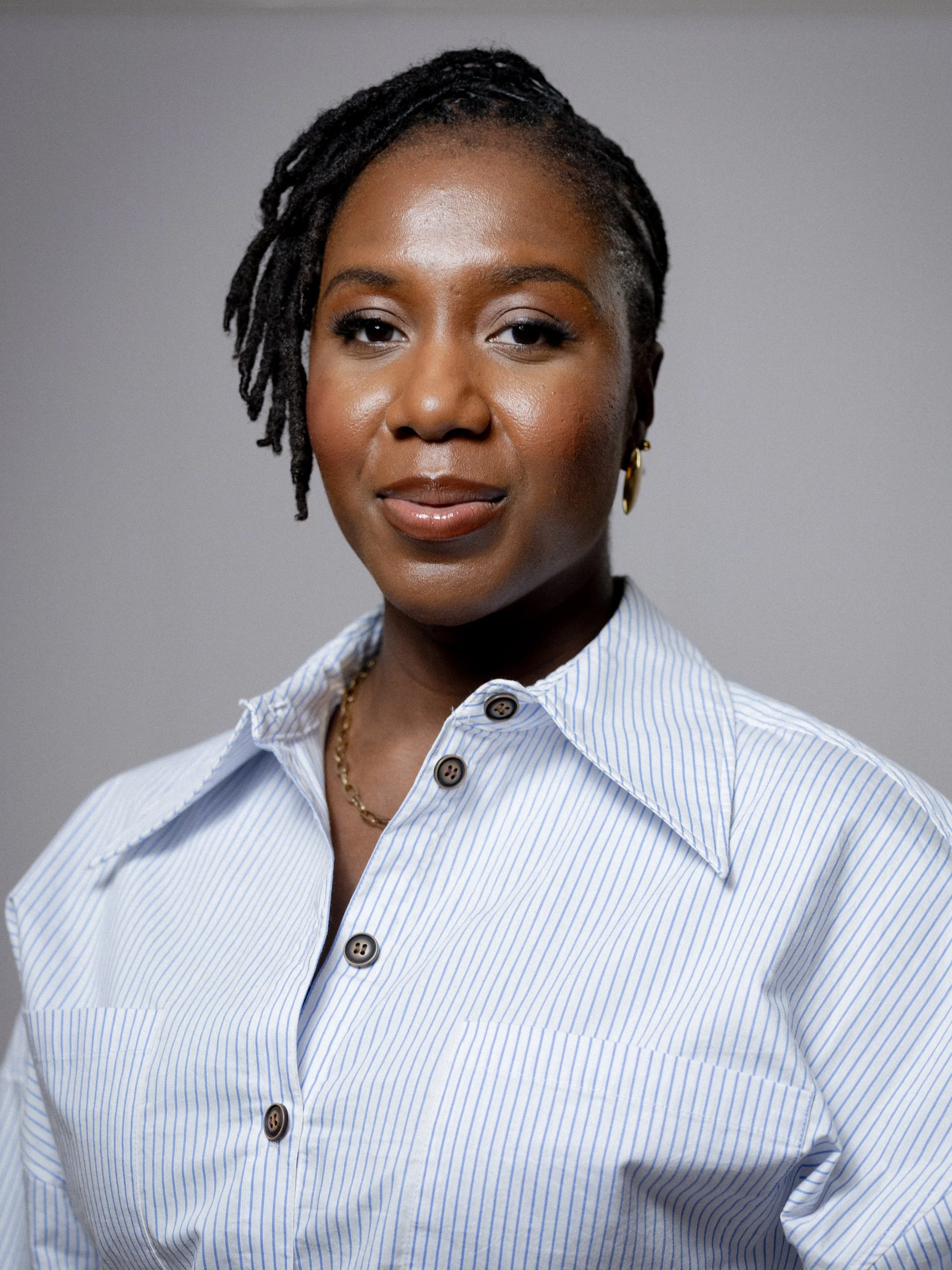 Close-up portrait of a Black woman with locs, wearing a blue-and-white striped shirt and gold earrings against a gray background.