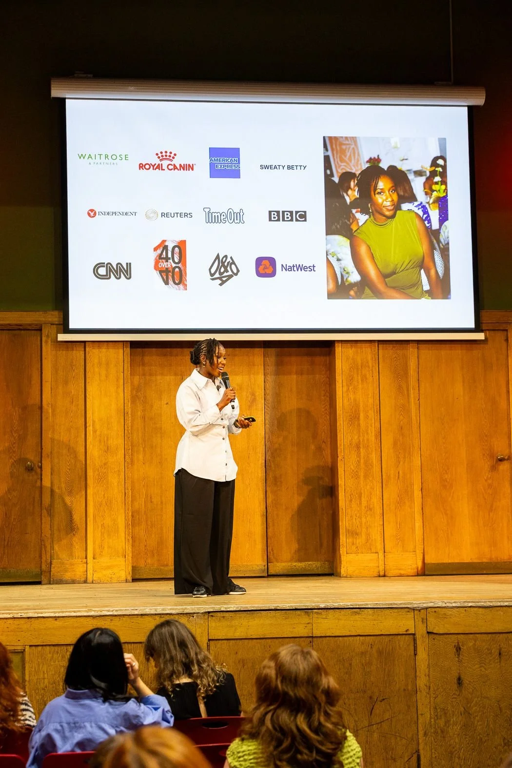 Woman speaking into a microphone onstage beneath a presentation slide showing corporate logos and a portrait, with an audience seated in the foreground