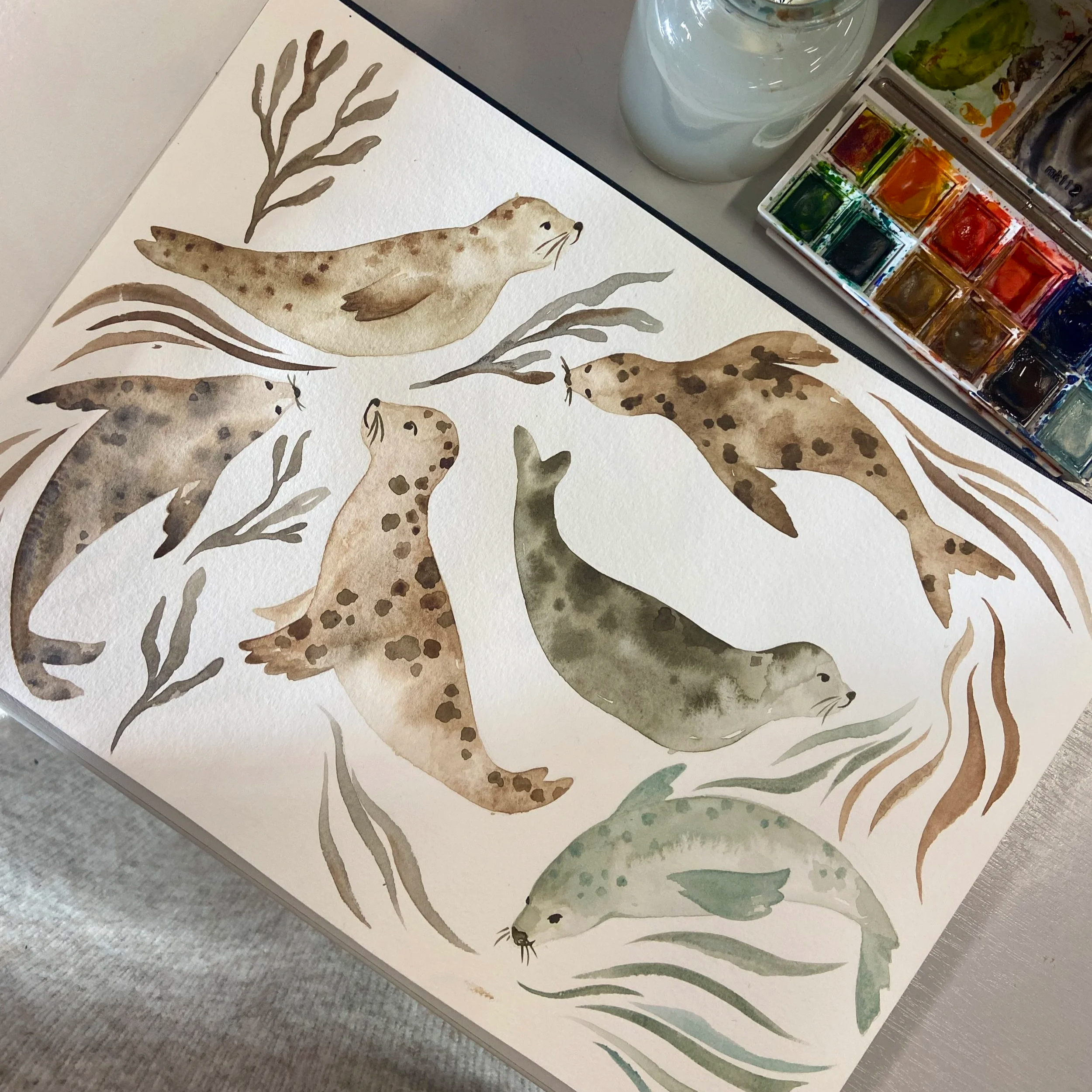 Watercolor painting of spotted seals swimming among seaweed, with a watercolor paint palette and water jar nearby. Hand painted surface print designs. Freelance print designer.