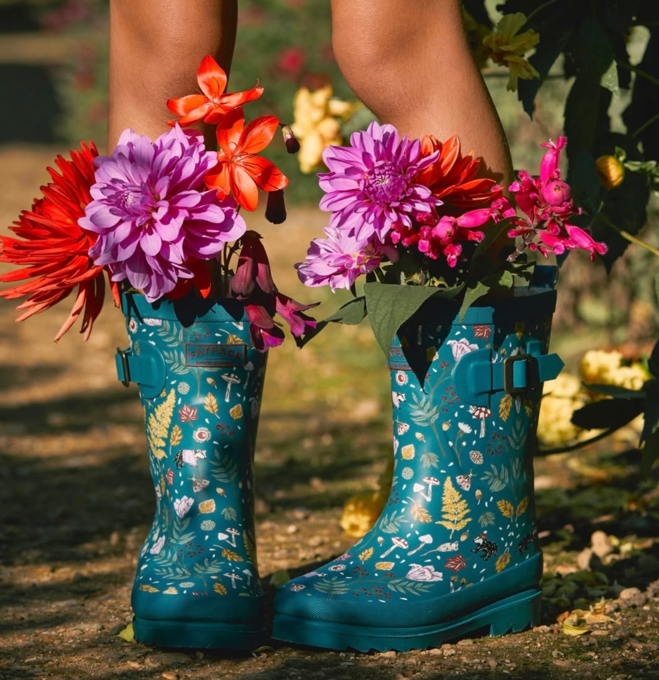 Person wearing teal floral-patterned rain boots filled with colorful flowers in a garden. Hand painted surface print designs. Freelance print designer.