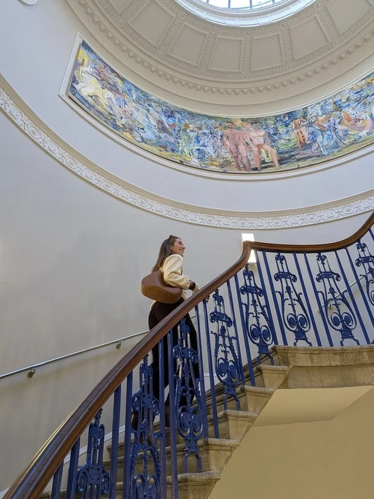 Woman climbing a grand staircase with ornate blue iron railings beneath a domed ceiling and colorful mural. Hand painted surface print designs. Freelance print designer.